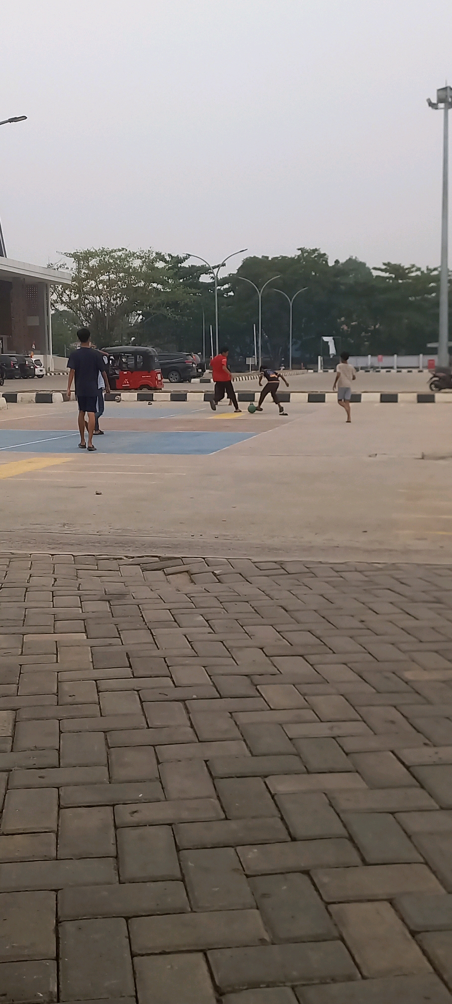 In Melayu, Indonesia, four young males are engaged in a game of soccer on an outdoor paved area. Two individuals in the foreground, one wearing a dark blue t-shirt and shorts, and another in a white t-shirt and grey shorts, are observed from behind, walking away from the camera. In the middle ground, two other males are actively playing soccer. One is wearing a red t-shirt and black pants, while the other is in a dark shirt and brown pants, kicking a soccer ball. In the background, a red auto-rickshaw and a dark-colored SUV are parked, with a building structure to the left and a line of trees and streetlights further back, suggesting an urban or suburban environment. The sky is overcast.