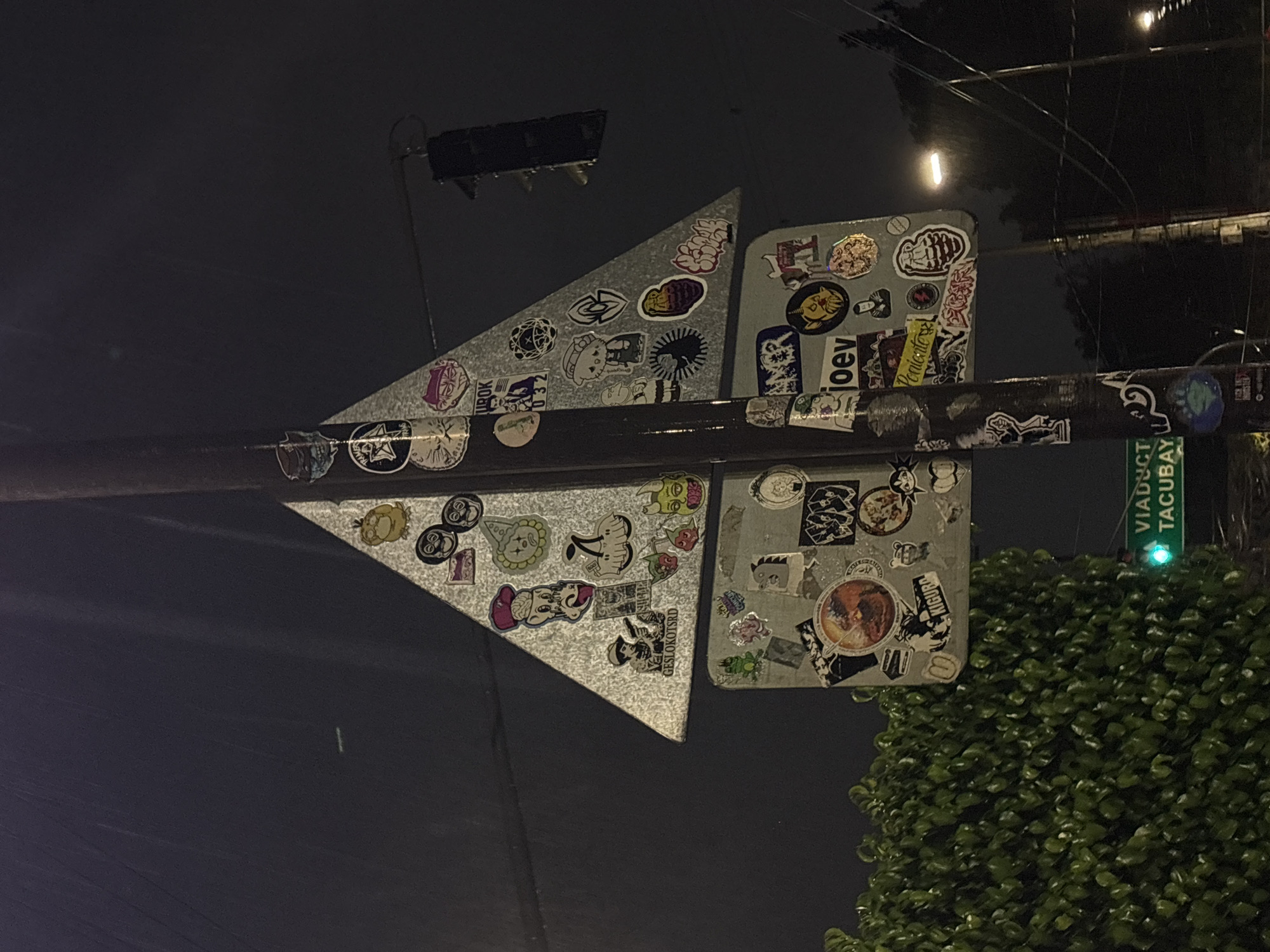 A street sign, heavily covered in stickers, is mounted on a horizontal pole. The sign consists of a pair of triangular plates pointing to the left and a pair of rectangular plates to the right. The pole is dark and also covered in stickers. In the background is a street sign showing "VIADUCT TACUBAYA" and a green light is illuminated next to it. A bush and some wires also appear in the background. The setting is an urban area at night in Mexico City, Mexico.