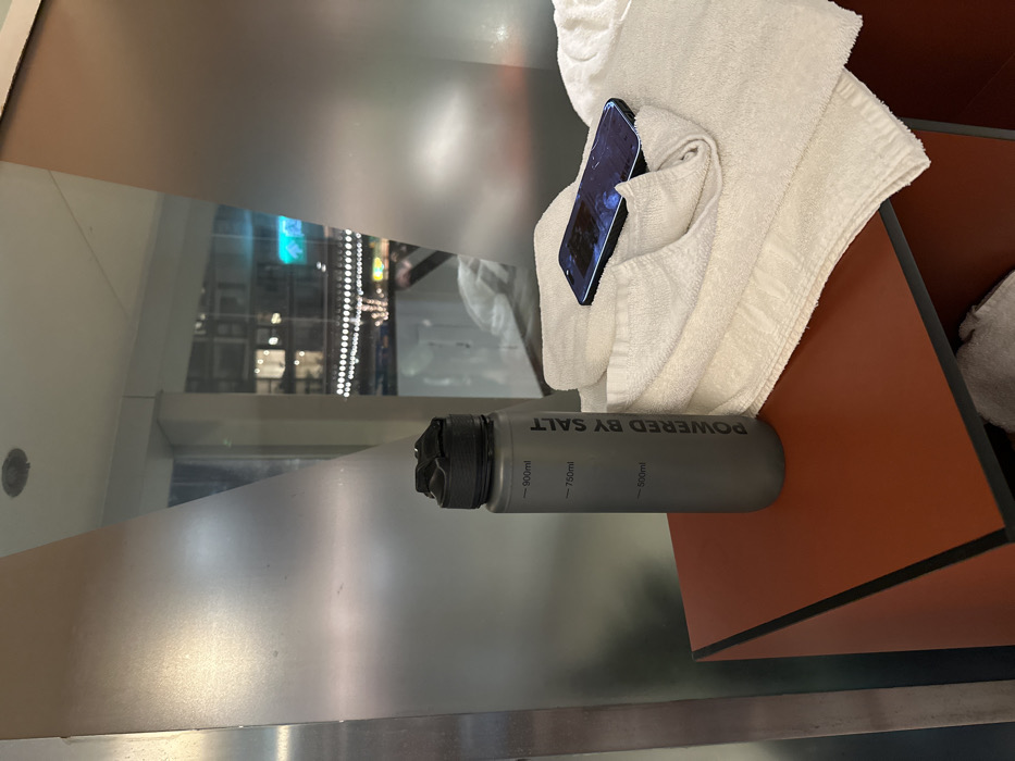 A gray water bottle with the words "POWERED BY SALT" and measurement indicators sits next to a folded white towel. An iPhone rests within the folds of the towel. A blurred reflection of a cityscape is visible in the shiny surface beneath the water bottle, indicating an indoor setting near a window. The items are placed on a table with brown and black accents.