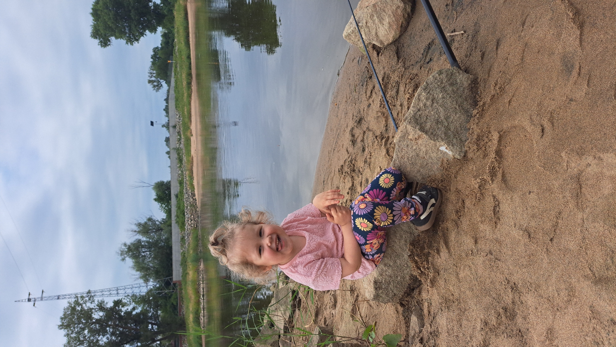 A young girl with blonde curly hair pulled into a high ponytail sits on rocks on a sandy riverbank. She is wearing a pink t-shirt with a floral pattern and colorful leggings with a floral design. Her tongue is sticking out, and she is smiling at the camera. The river is calm and reflects the cloudy sky. In the background, trees and a power line tower are visible. A fishing rod is resting on the sand to the right of the girl. The scene appears to be a park or natural area within the city of Krosno Odrzańskie, Poland, likely near the Oder River.