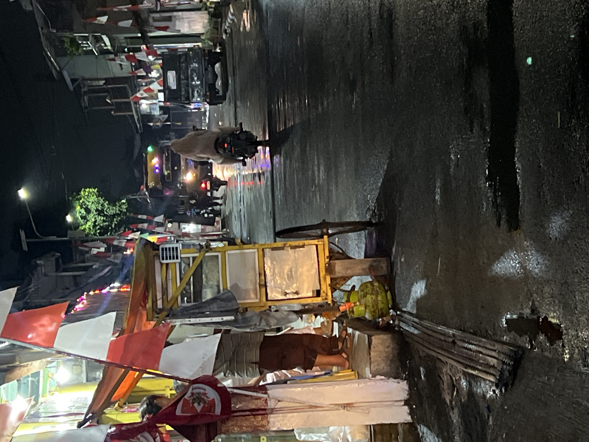 The image depicts a nighttime street scene in Citeureup, Indonesia. The street is wet and reflects the lights of vehicles and surrounding buildings. In the foreground, a vendor's cart, painted yellow and white, is parked on the side of the road. A large, green gas cylinder sits at the base of the cart, and a stack of metal rods lies on the wet pavement next to it. Red and white flags, likely Indonesian flags, are displayed prominently on and around the cart, suggesting a local celebration or event.
Several people are present. A person stands near the cart, partially obscured by a draped fabric and a flag. Further down the street, a person is riding a motorcycle, wearing a light-colored hijab. Other individuals are seen on motorcycles and walking in the background. Vehicles, including a truck and a bus, are visible further down the street. The buildings lining the street are multi-story structures, some with windows and balconies. The overall atmosphere is that of a bustling urban environment at night, possibly during a festive occasion, given the presence of flags and the nighttime activity.
