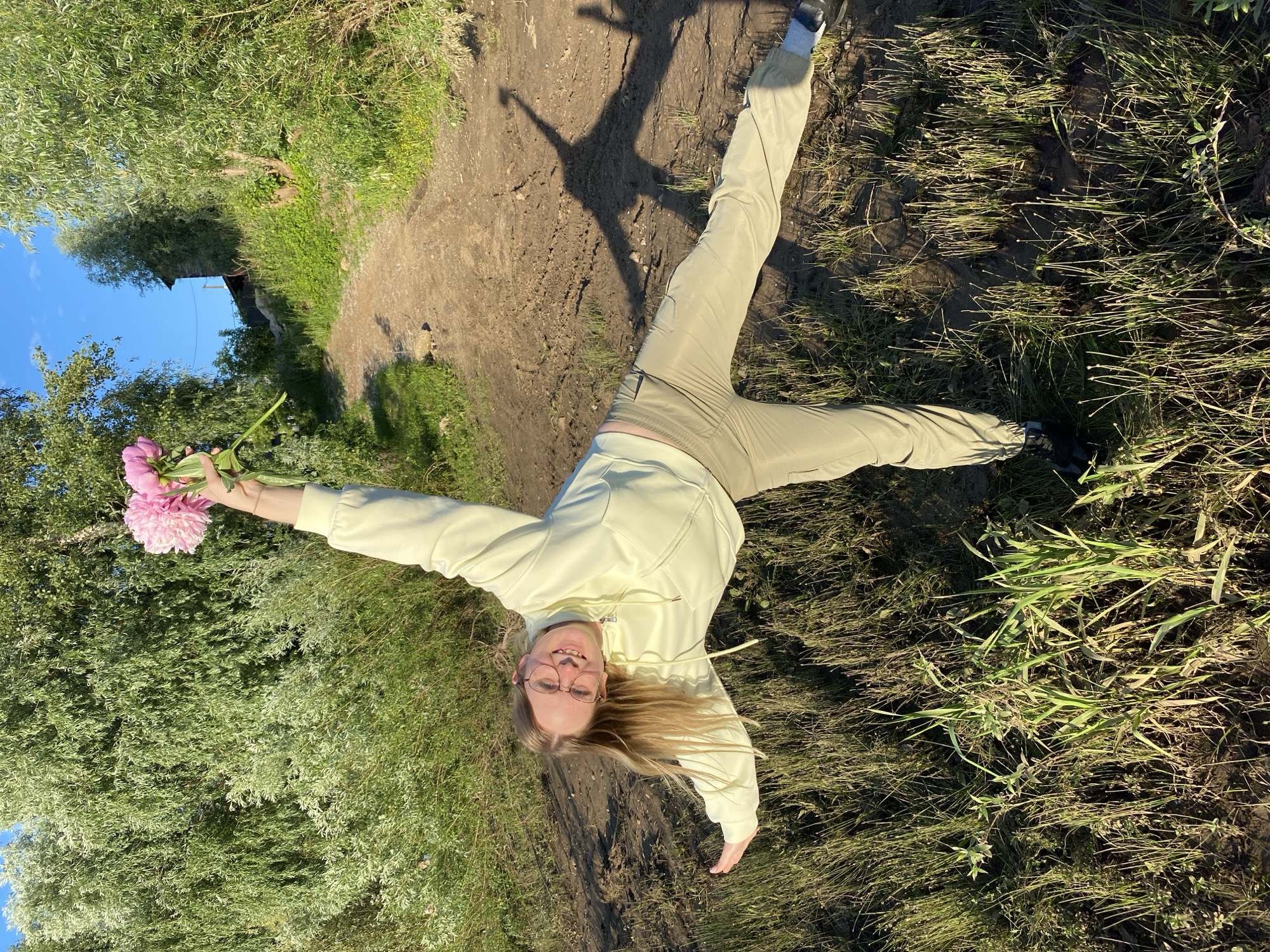 A young woman with blonde hair and glasses lies on her back on a dirt path in an outdoor setting. She is wearing a light yellow hoodie and light khaki pants. Her arms are outstretched, and she is holding a bouquet of pink flowers in her left hand. The sun is shining, casting a shadow of the woman on the path, suggesting it is daytime. She appears happy and relaxed. The scene is surrounded by green grass and trees, with a clear blue sky visible. The image was taken in Chaikovsky, Russia.