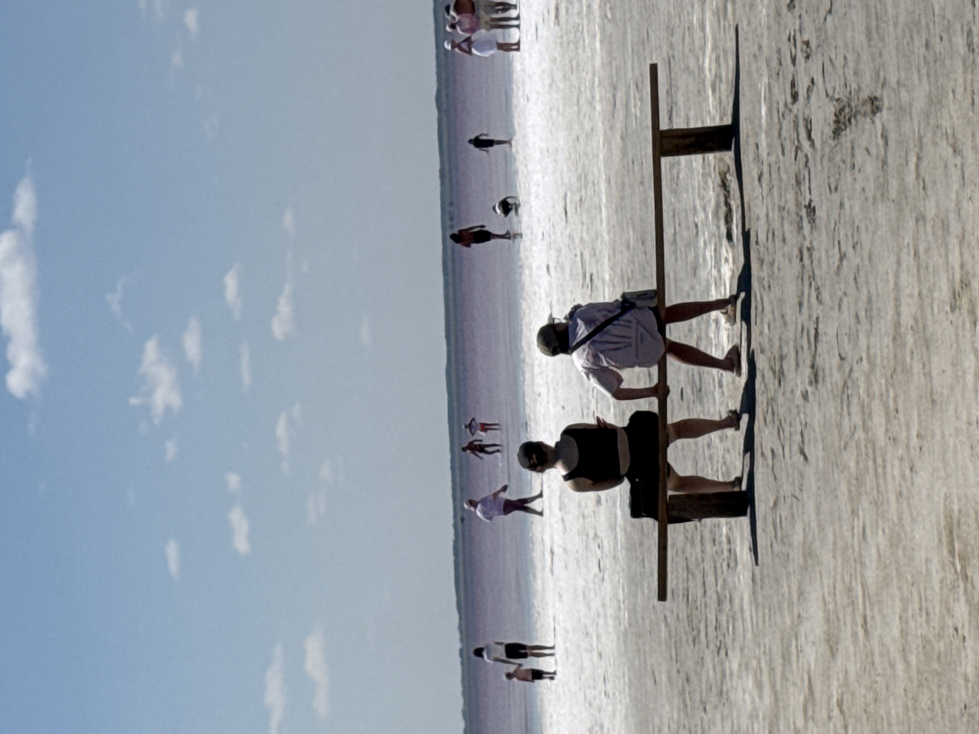 The image depicts a sunny day at the salt flats of Бурсоль, Russia. Two individuals are seated on a wooden bench in the foreground, facing away from the camera. The person on the left is wearing a black tank top and shorts, while the person on the right is wearing a light-colored shirt, shorts, and a cap.

In the background, several other people are visible in the distance, walking on the salt flats and in the shallow pink-colored water. The environment is characterized by a vast expanse of white salt crystals leading to a body of water that appears to be a salt lake. The sky is blue with scattered white clouds. The overall scene conveys a sense of leisure and recreation in a unique natural landscape.