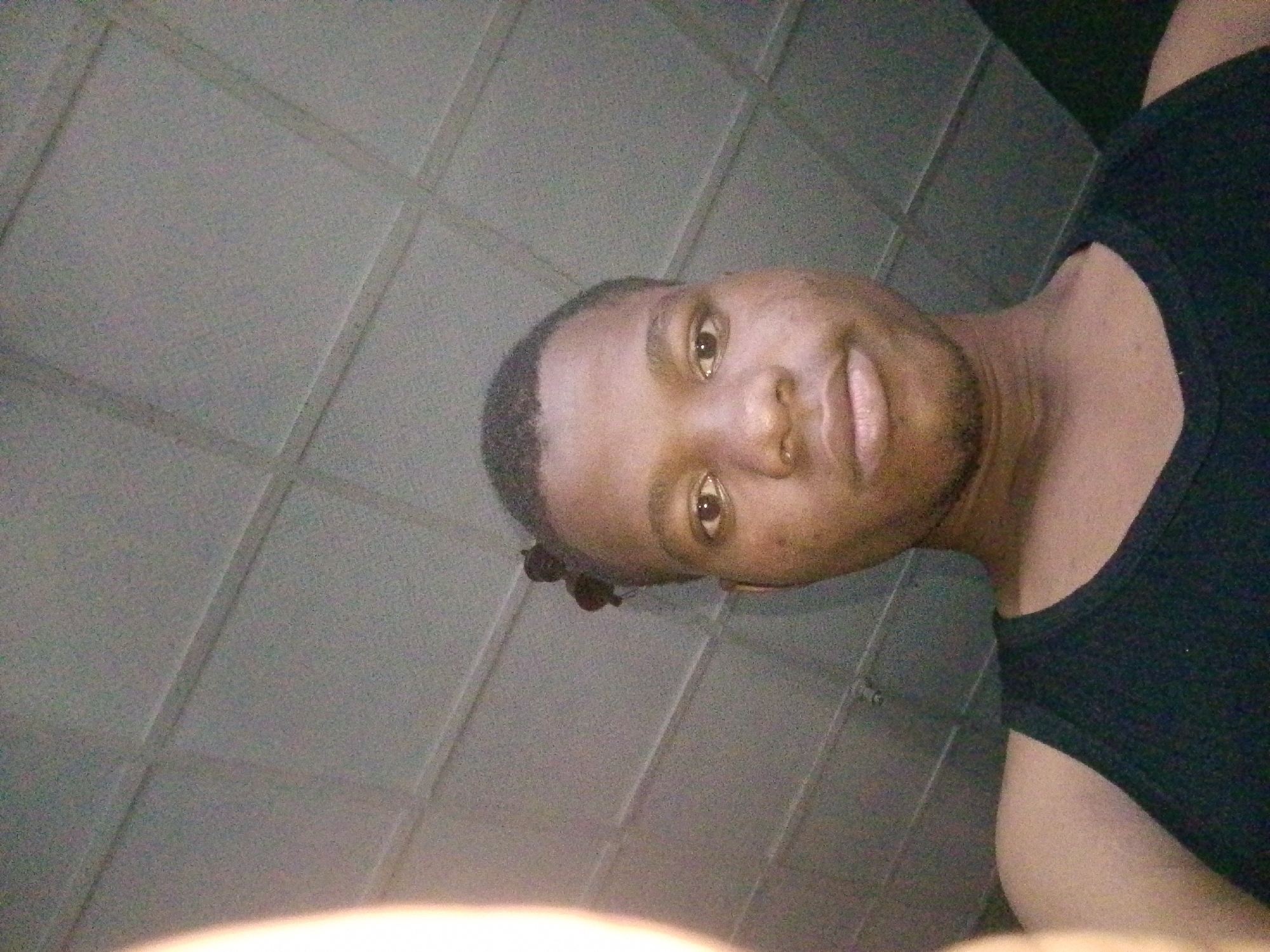 This is a low-angle, waist-up, selfie-style photograph of a young Black man in Ipetumodu, Nigeria. He is wearing a black tank top. His head is tilted to his left, and he is looking directly at the camera. His facial expression appears neutral. The background consists of a tiled wall with a grid pattern. The lighting suggests it is daytime, possibly indoors due to the uniform lighting and lack of shadows. The overall mood is casual and personal.