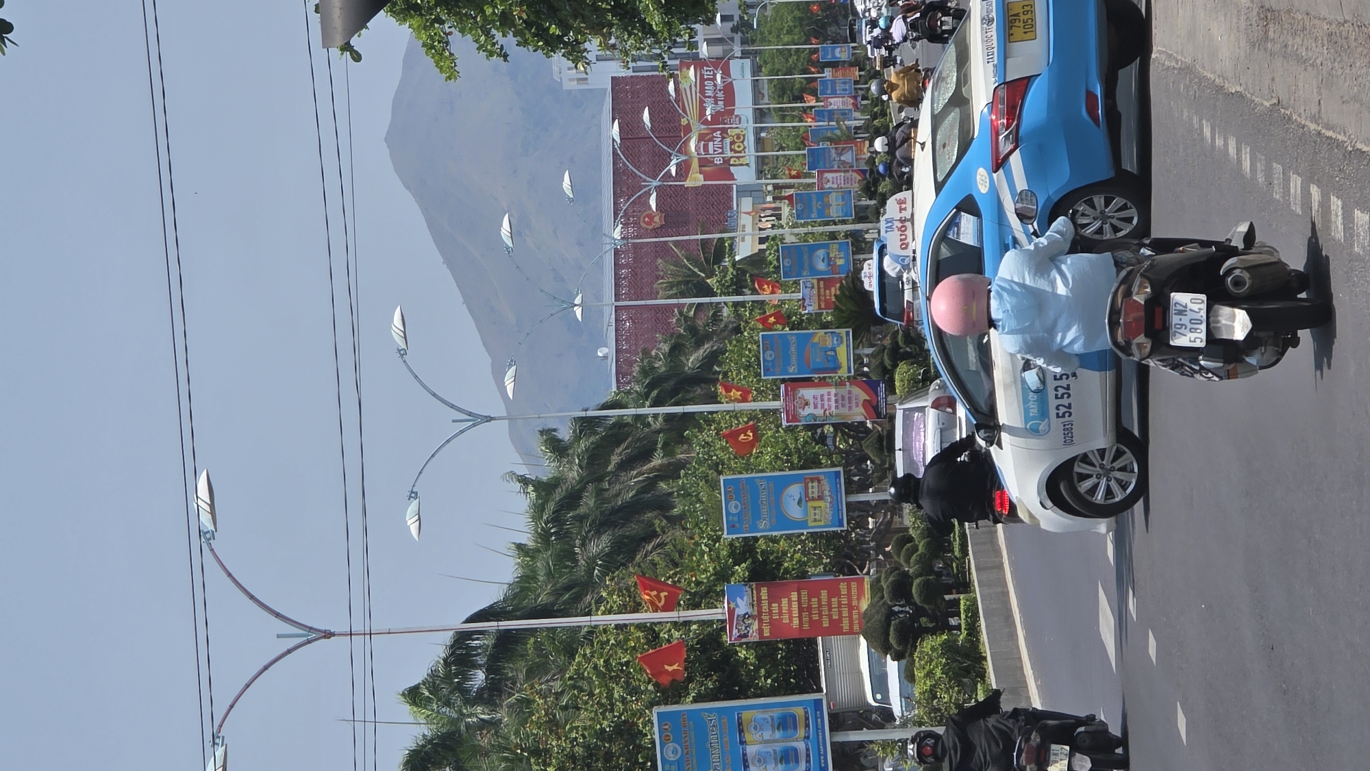 The image depicts a street scene in Nha Trang, Vietnam, with several billboards and Vietnamese flags lining the roadside. A mountain range is visible in the background. In the foreground, a white and blue taxi with "TAXI QUỐC TẾ" (International Taxi) and a phone number (02583) 52 52 5... printed on its side is stopped in traffic. A person wearing a pink helmet and light blue shirt is on a black motorbike next to the taxi. The motorbike's license plate number is 79-N2 580.40. Several other motorbikes and vehicles can be seen further down the street. Banners wishing a Happy New Year and advertising the BIVINA brand are noticeable. Additional advertisements featuring "Samcanest" and "Anvinest" are also displayed. Another sign mentions the anniversary of the liberation of Khánh Hòa province (4/1975 - 4/2026) and the reunification of Vietnam (30/4/1975 - 30/4/2026). Streetlights with leaf-like designs are visible along the road.