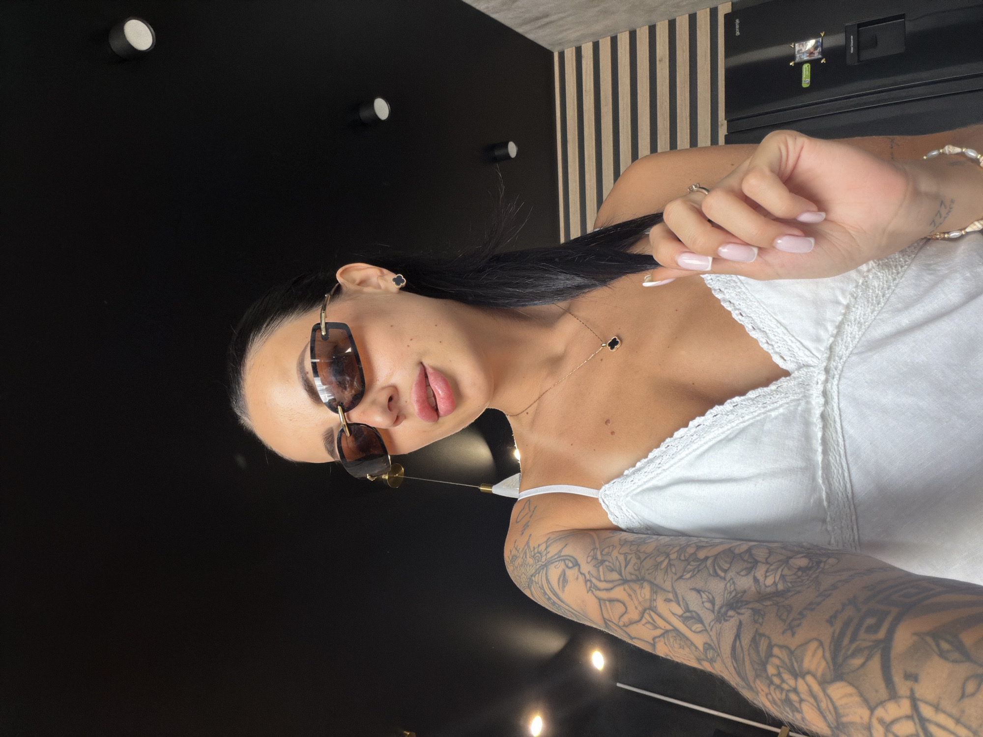A woman wearing sunglasses and a white camisole is posing for a selfie in an indoor setting. Her left arm is covered in tattoos, and she is holding a strand of her dark hair with her right hand. She is also wearing a necklace with a clover-shaped pendant and small clover earrings. The background includes a black ceiling with recessed lighting and a wooden slatted wall. A black refrigerator is visible on the right side of the image. The image is taken from a low angle, looking up at the woman.