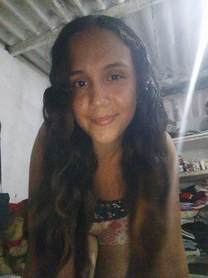 A woman with long, dark wavy hair smiles at the camera. She wears a patterned tank top. The background shows a cluttered room with items stacked on shelves or furniture. The lighting is uneven, with a bright spot on the ceiling. A teddy bear is visible in the lower left corner. The room appears to be modest, possibly a bedroom.