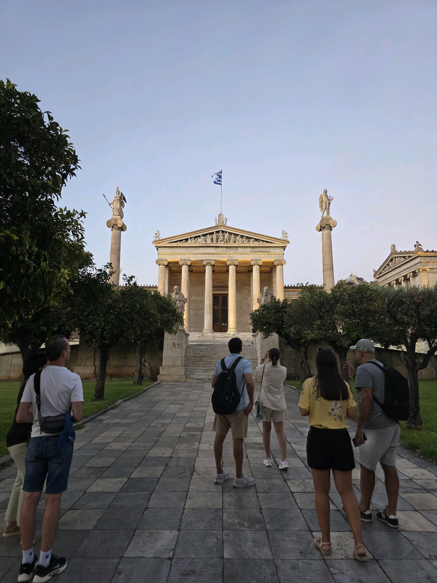 The image depicts the Academy of Athens in Greece, a prominent neoclassical building situated in the city center. In the foreground, a group of five individuals, likely tourists, are observed from behind, walking on a paved walkway. The group comprises two men, two women, and one person whose gender is not discernible. They are dressed in casual attire suitable for warm weather, with some carrying backpacks.
The Academy building dominates the background, characterized by its grand architecture featuring classical columns, pediments adorned with sculptures, and a Greek flag waving from a flagpole atop its central structure. Two imposing statues on tall pedestals flank the main facade, adding to the monumental feel of the scene. Trees with lush green foliage are planted along the walkway, providing a natural element to the urban landscape. The sky above is clear, with a pale blue hue, suggesting it is either early morning or late evening. The overall atmosphere is serene, with the focus on the architectural grandeur and the presence of visitors.