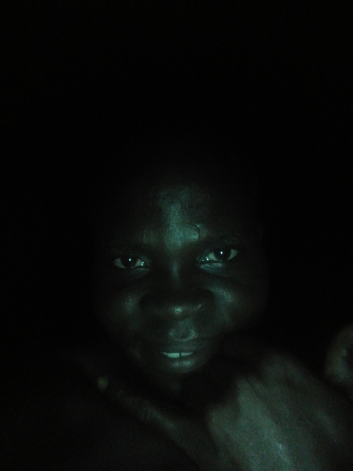 A dimly lit, close-up image shows the face of a person with dark skin. The person is smiling slightly, and their eyes are visible. A hand or arm is partially visible in the lower right corner of the frame. The background is dark and indistinct. There is no information about the location in Ikorodu, Nigeria.