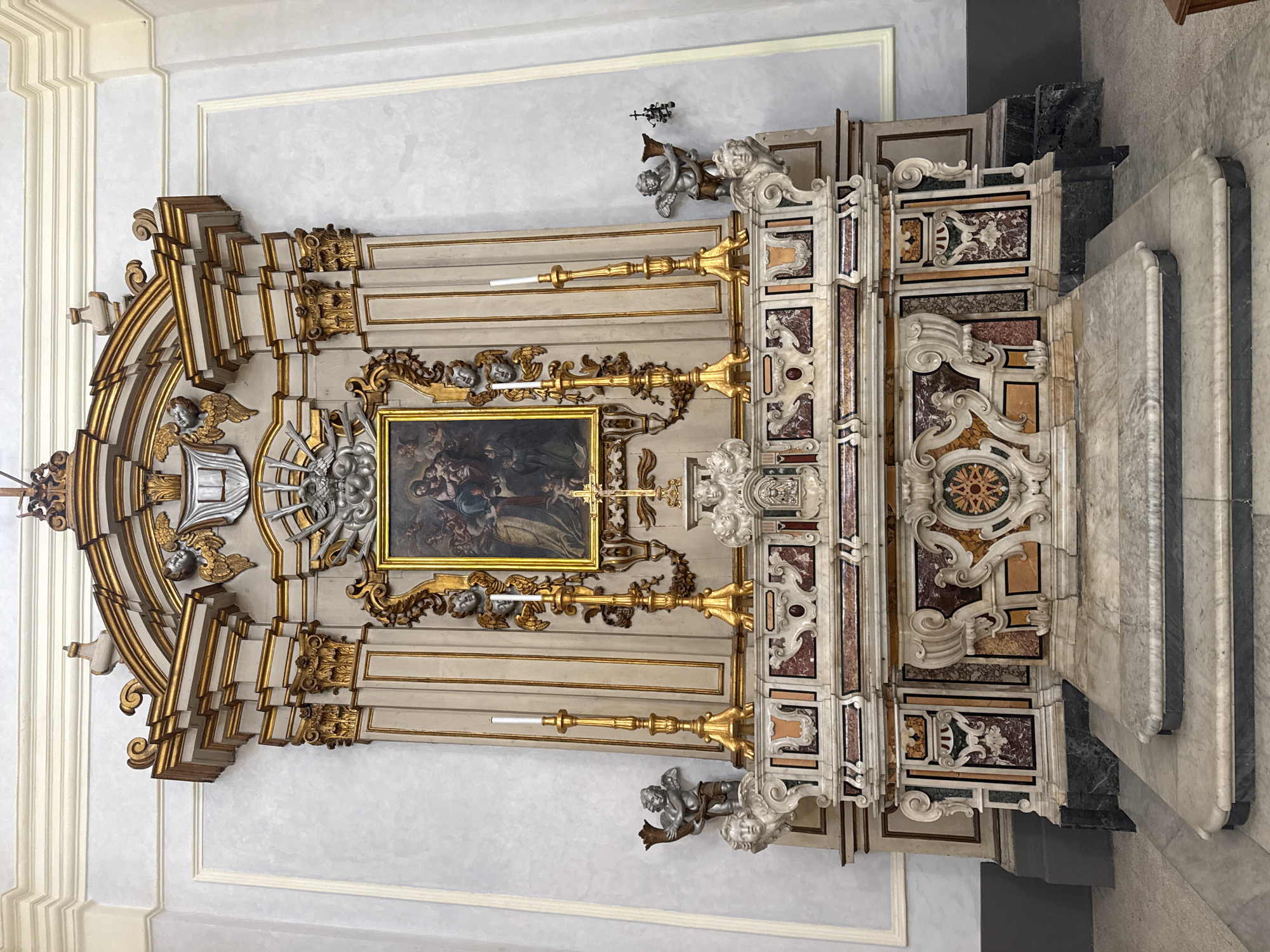 This full-length, slightly tilted indoor photograph captures an ornate Baroque or Rococo-style altar, likely within a church in Monopoli, Italy. The elaborate altar is composed of white and gold painted wooden elements combined with richly inlaid marble (pietra dura or scagliola) panels featuring various colors such as red, brown, black, beige, and green, arranged in intricate scrollwork and geometric patterns.
At the center of the altar is a rectangular painting, set within a gold frame, depicting a religious scene with multiple figures, rendered in somewhat dark tones. Flanking the painting and integrated into the altar's structure are several large, golden candlesticks holding white candles. Numerous sculptural elements, including white and silver-toned putti or angels, adorn the upper and lower sections of the altar, contributing to its grand and opulent appearance. Some angels appear to be supporting elements or decorative motifs.
To the right of the central altar structure, there are architectural elements constructed from light and dark gray marble, including what appears to be steps or a side altar. The altar is set against a light gray wall that features subtle framed paneling. The overall impression is one of rich detail, luxurious materials, and intricate craftsmanship, characteristic of historical ecclesiastical art and architecture found in Italy. There is no discernible activity taking place, and no visible text in the image. The lighting is consistent with an indoor setting, likely diffused or artificial.