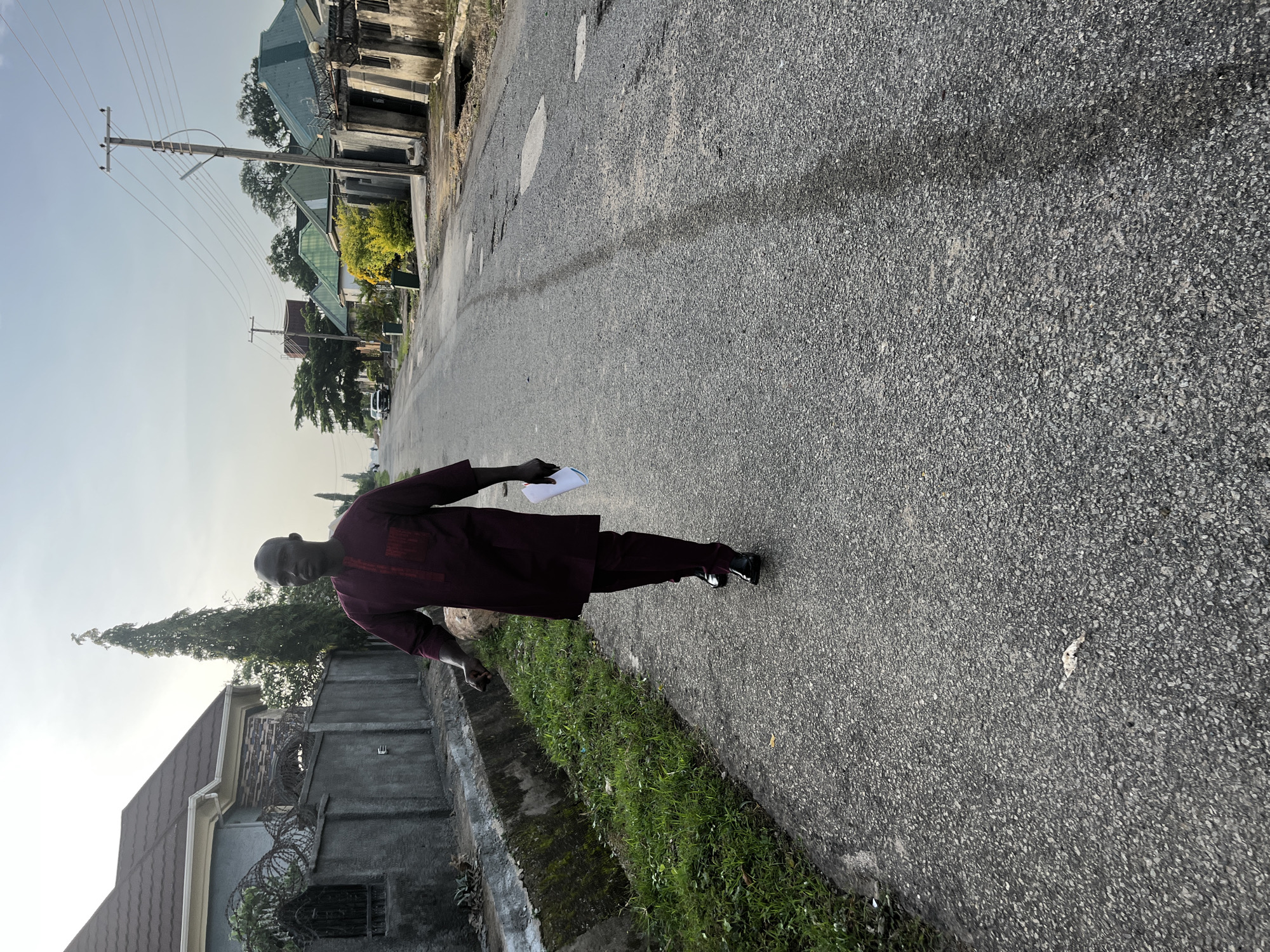 A man in a maroon traditional outfit walks along a paved road in Abuja, Nigeria. He is holding a white piece of paper in his right hand. The road is bordered by a concrete drainage ditch on the left, with green grass growing within it. Beyond the ditch, there is a grey concrete wall topped with barbed wire, and a modern house with a brown tiled roof. Several other houses with green roofs are visible further down the street, along with utility poles and overhead wires. The sky is overcast, with the sun casting a soft light. The overall scene suggests a residential area in Abuja.
