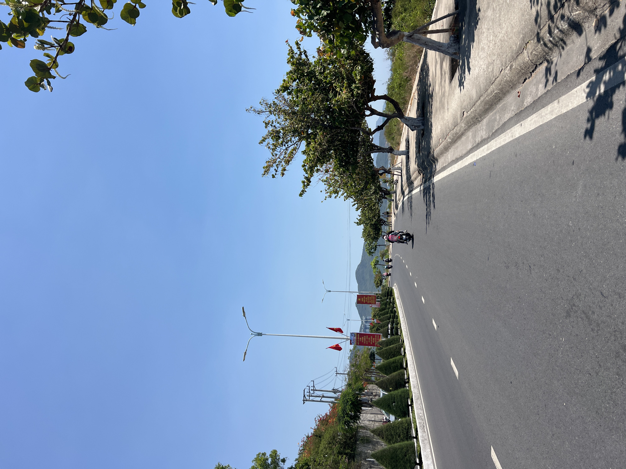 The image captures a road in Bắc Nha Trang, Vietnam, on a sunny day. The road stretches into the distance, flanked by trees on one side and a landscaped area with flags and light poles on the other. Motorbikes are visible on the road. The sky is clear blue.