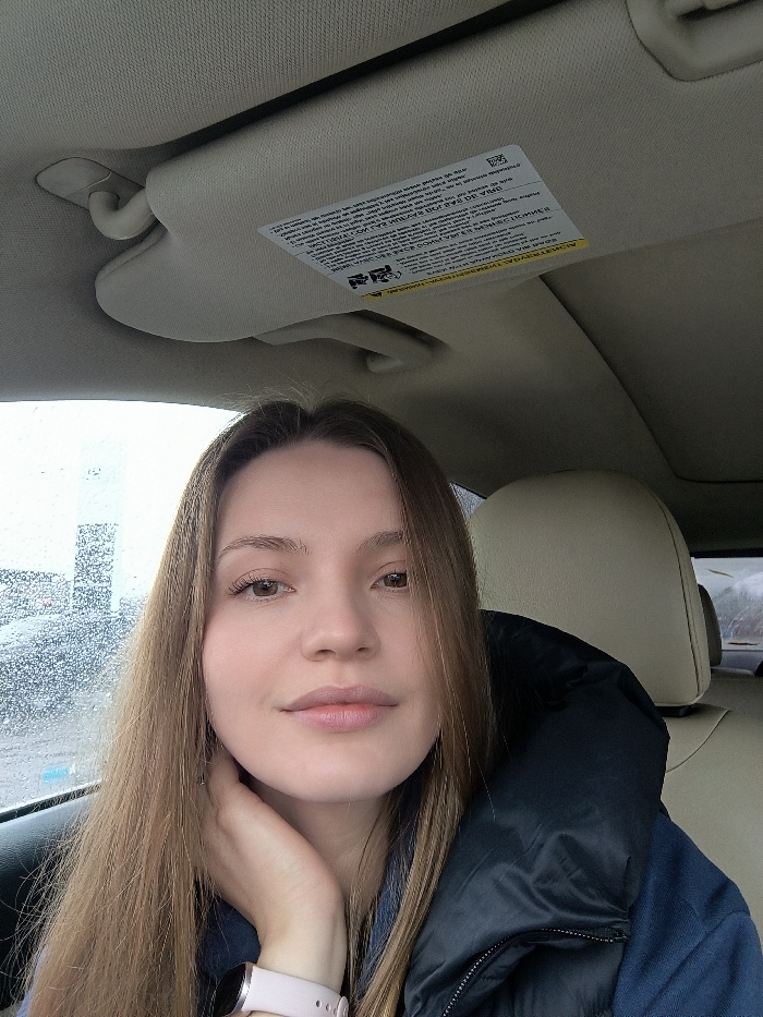 The image displays a young adult female seated inside a vehicle, in what is stated to be the city of Illintsi, Ukraine. No specific landmarks are visible in the image to independently verify this geographic location.
**Subject:**
A female with long, straight, light brown hair and brown eyes is positioned in the foreground, looking directly at the camera with a neutral expression and a slight closed-lip smile. Her left hand is raised, resting with fingers near her jawline/neck. She wears a light pink smartwatch with a metallic face on her left wrist. Her attire includes a dark-colored (possibly black or dark blue) puffy jacket worn over a blue shirt. The subject appears to be in the driver's seat.
**Vehicle Interior:**
The vehicle's interior features light-colored (beige or cream) upholstery on the headliner, sun visor, and the visible portion of the passenger seat headrest. A warning label, affixed to the sun visor above the subject's head, contains multi-lingual text.
**Exterior Environment:**
Through the driver's side window, the view is partially obscured by water droplets on the glass, indicating rain or moisture. Distant, indistinct vertical structures are visible through the window, suggesting an urban or industrial background under an overcast sky.