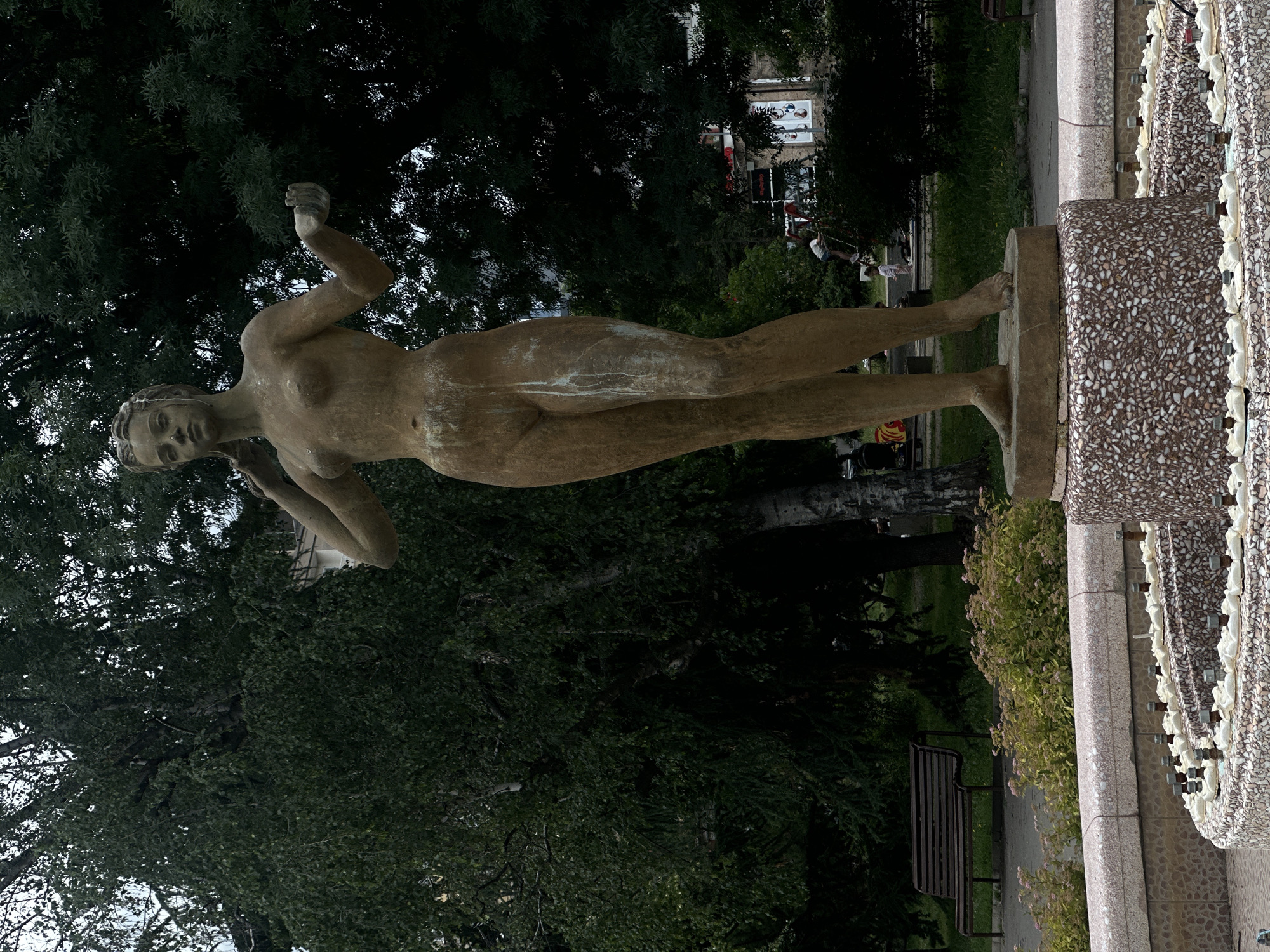 This is a full-body, eye-level shot of a weathered, nude female statue standing on a circular base. The statue is made of a light-colored material, possibly bronze or stone, and shows signs of age and wear, with some green and white streaks running down its body. The woman has her left arm raised to her head, and her right arm is extended outwards with her palm facing up. She has a serene expression on her face and her hair is styled in a bob. She stands in what appears to be a public park or square in Sofia, Bulgaria, as indicated by the presence of trees, foliage, a park bench in the background, and the fountain structure in the foreground. The fountain base is made of a mosaic of small, speckled stones. In the background, behind the statue and trees, parts of buildings and a street can be seen. Two children are visible in the mid-ground, one on a playground structure and another standing nearby. The overall atmosphere is somewhat somber due to the overcast sky and the aged appearance of the statue.