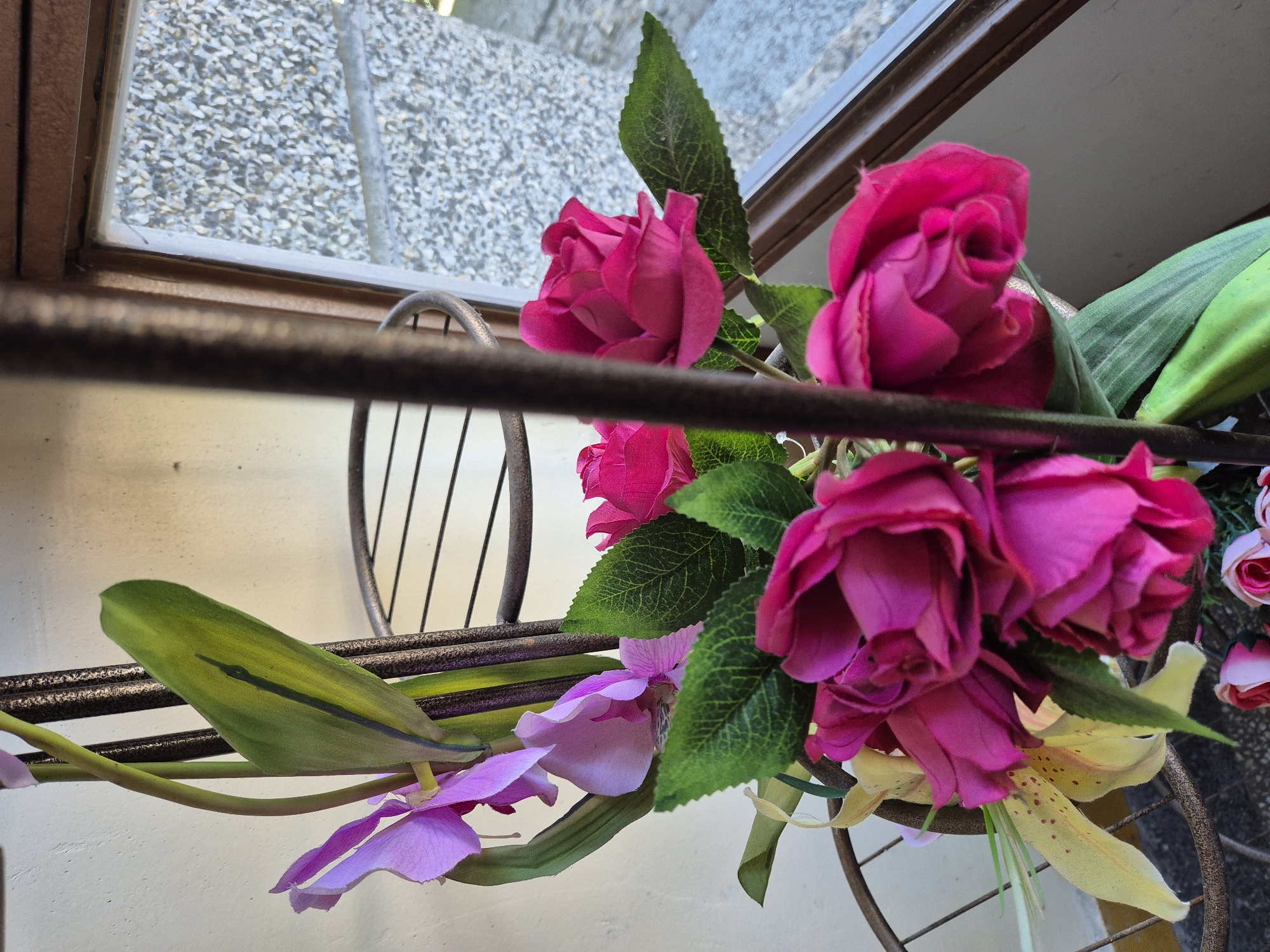 A close-up, medium shot shows a floral arrangement of artificial flowers on a metal stand. The arrangement includes several bright pink roses and some purple orchids. There are also some green leaves and a yellow lily with red speckles. The floral stand is visible, with a dark brown metal frame. Behind the flowers, a window is partially visible, with a textured grey wall on the outside. The background is a light-colored wall. The image is taken from a slightly low angle, looking up at the flowers.