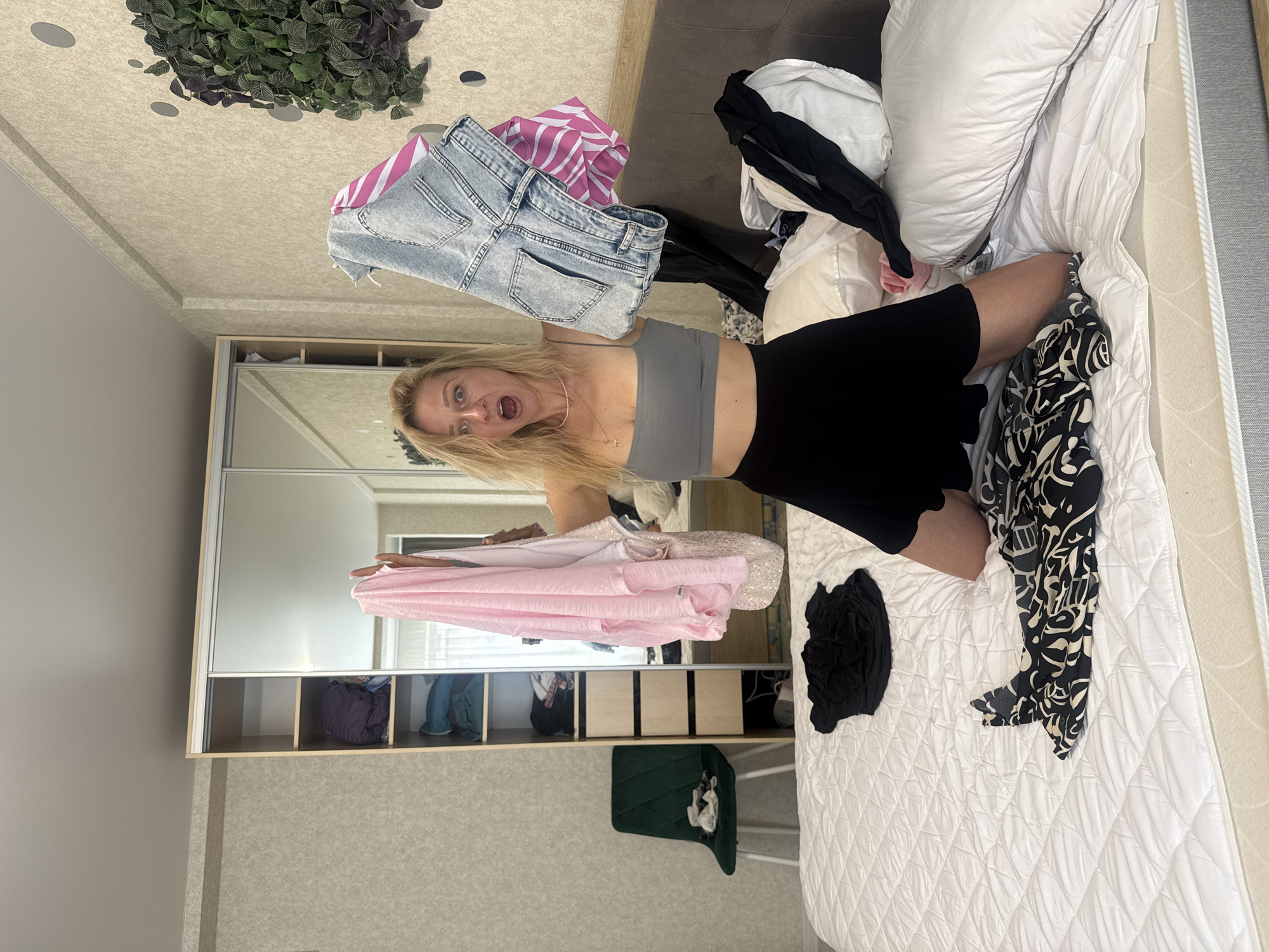 A woman kneels on a bed in a bedroom. She is wearing a grey crop top and a black skirt. Her mouth is open in surprise as she holds up a pile of clothes in her hands. The bed is covered with clothes, and there is a wardrobe and a chair in the background. The room appears to be in disarray.