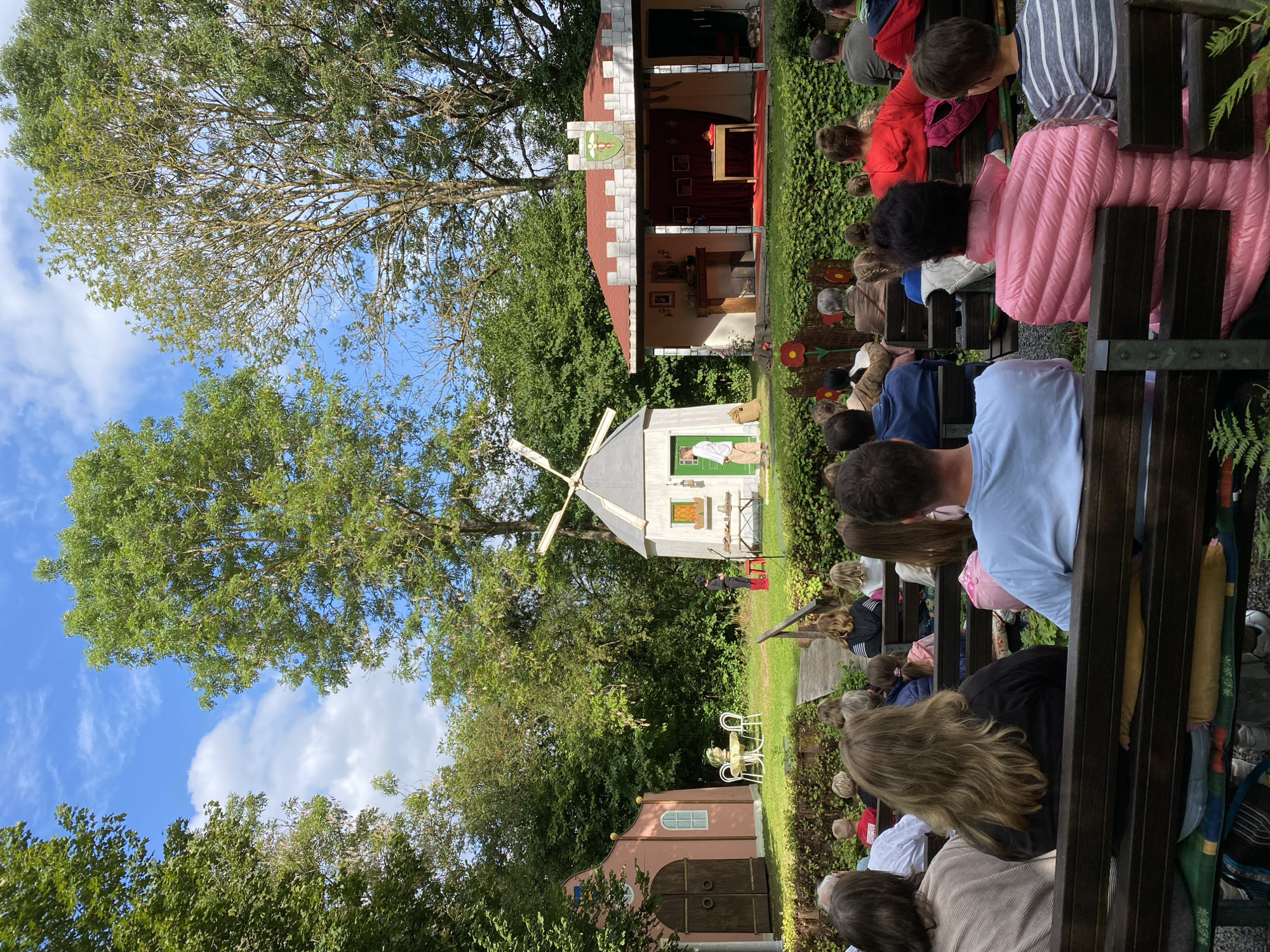 In an outdoor theater setting, an audience sits on wooden benches arranged in rows, facing a stage. The stage is decorated with a small pink building to the left, resembling a house, and a windmill structure to the right. In the center of the stage, a small castle-like facade with battlements stands. Two actors are on stage, one standing near the windmill, and another near the castle structure. The backdrop consists of lush green trees, and the sky above is blue with scattered clouds. The location is the Freilichtbühne in Flammersfeld, Germany, a well-known open-air theater.