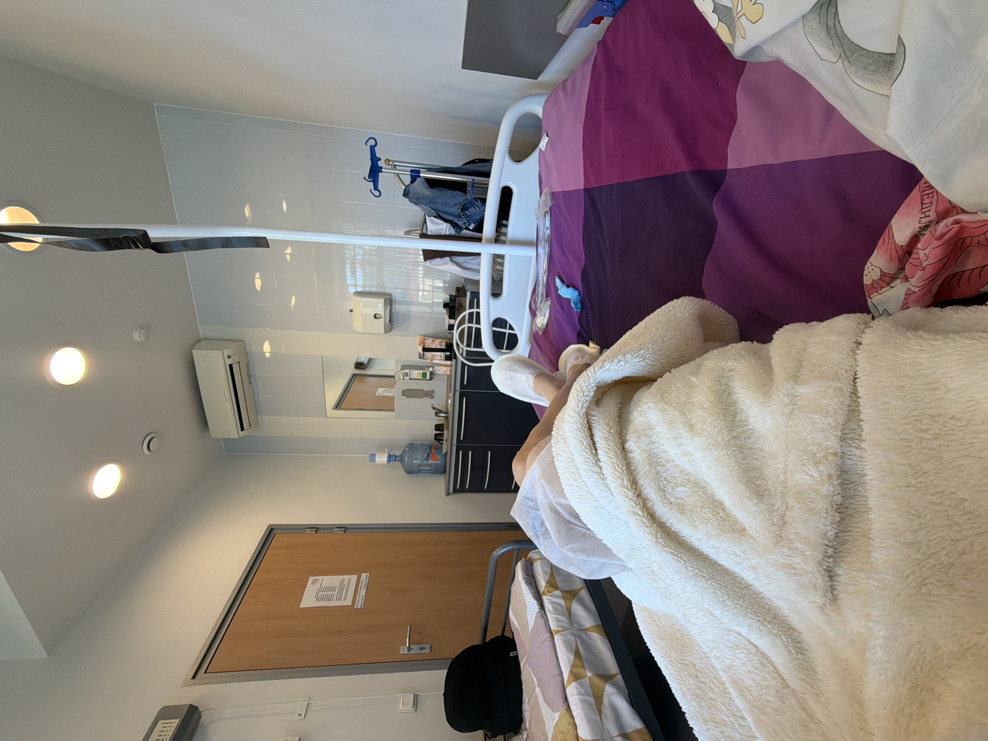 The image depicts what appears to be a patient's room in a medical facility, possibly in Saint Petersburg, Russia, though explicit confirmation of the location is absent. A hospital bed occupies a significant portion of the frame, covered with multicolored bedding and a thick, off-white blanket. An individual, presumably the patient, is partially visible under the blanket, wearing white socks.
Medical equipment is present, including an IV pole and a water dispenser on a counter near a sink and mirror. The room itself is sterile with tiled walls. A door with informational notices on it is also visible.