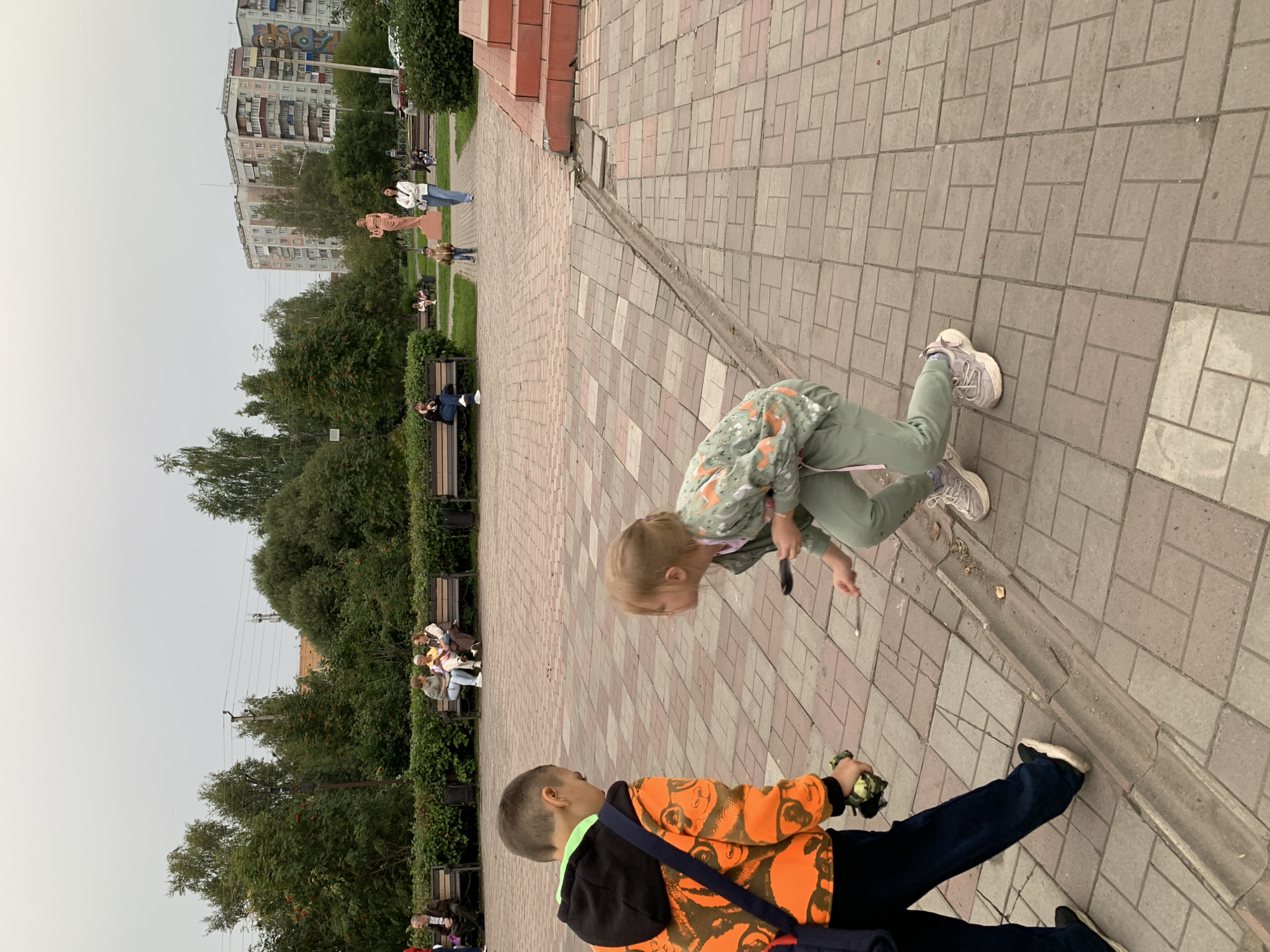In Prokopyevsk, Russia, two young children are observed in a public outdoor setting. In the foreground, a boy wearing an orange hooded jacket with a panda print and dark pants is walking away from the camera. He is carrying a green object in his left hand. To his right and slightly in front, a younger girl in a green patterned jacket and green pants is crouched down, holding a small stick and appearing to interact with the ground. The children are on a paved walkway made of interlocking brick-like stones, with a drainage channel running alongside it. In the midground, several benches are occupied by adults, some engaged in conversation. A large statue of a woman is visible in the background, flanked by trees and residential buildings. The sky is overcast.