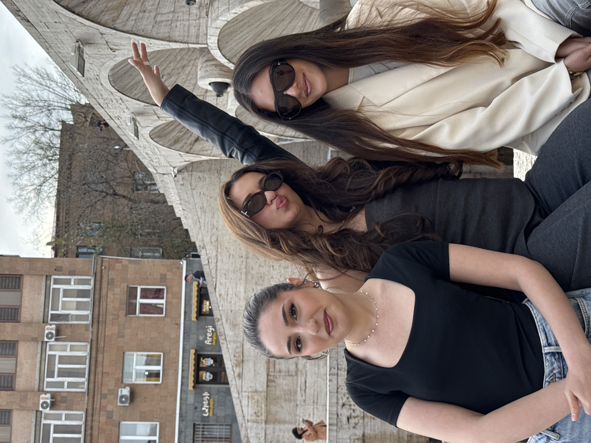 Three young women are posing for a photograph. The women are standing in front of the Cascade complex in Yerevan, Armenia. The woman in the foreground is wearing a black top, a silver necklace, and light blue jeans. The second woman is wearing a black leather jacket and sunglasses. The third woman is wearing a cream-colored blazer and sunglasses. The woman in the black leather jacket has one arm raised. In the background, there is a beige-colored building with windows and air conditioning units. A business with signage is also visible.