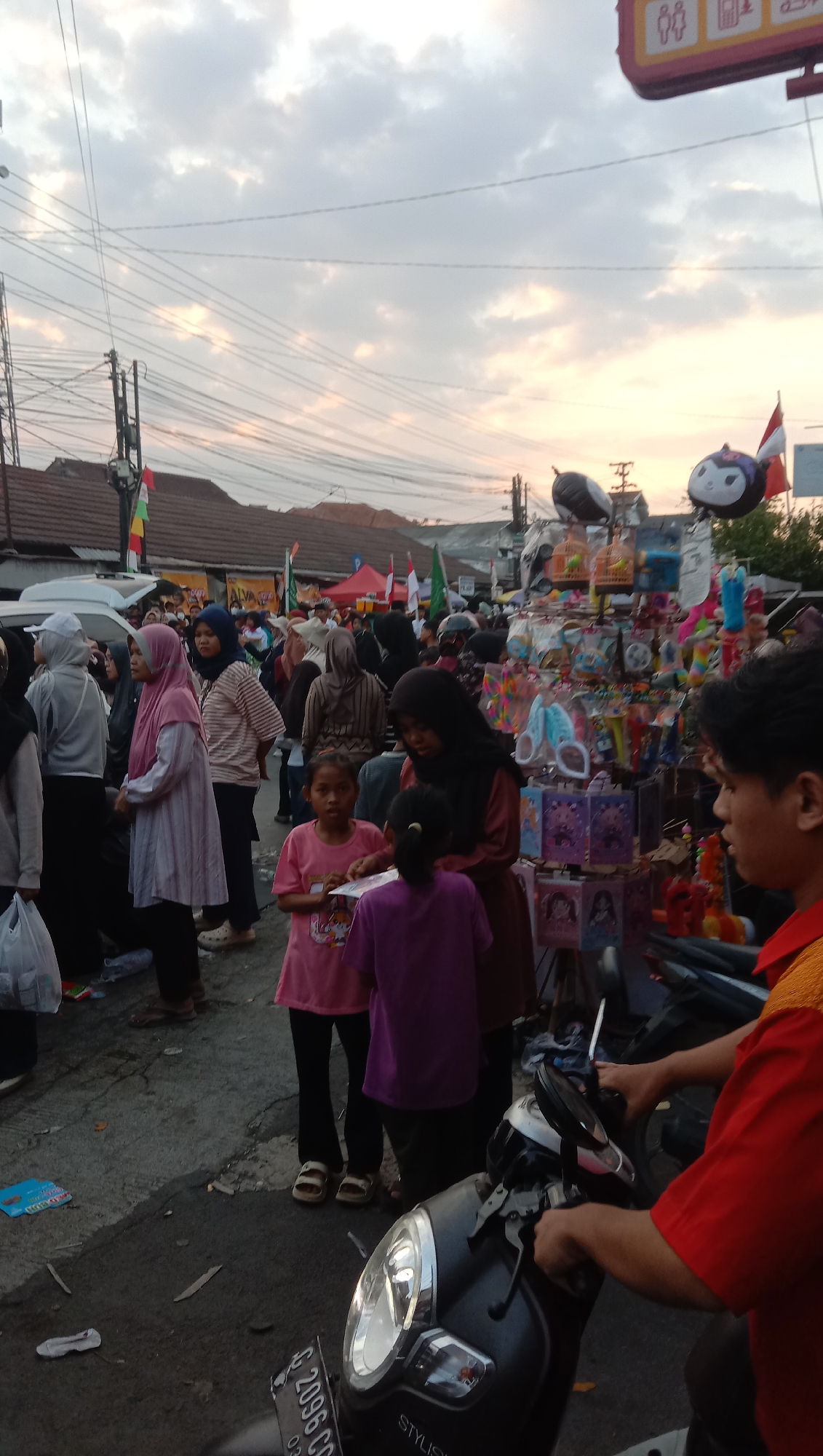 The image depicts an outdoor street market scene at dusk in Jatisawit, Indonesia. The sky is filled with soft clouds, illuminated by the setting sun, casting a warm glow. The foreground is dominated by a dark-colored scooter, with a person in a red shirt seated on it, holding the handlebars. A license plate on the scooter reads "G 2096 CD".
The street is paved and somewhat littered with small pieces of debris. Several people are present, mostly wearing casual clothing and headscarves. In the mid-ground, two young girls are standing near a stall. One girl, wearing a pink t-shirt, is looking towards the viewer. The other, in a purple t-shirt, is partially obscured. Behind them, a woman in a dark headscarf and a brown dress is standing.
Further back, a variety of market stalls are visible. One stall features colorful balloons, including two with cartoonish faces, and other small trinkets displayed on shelves. More people are browsing at these stalls, and tents are set up in the background, suggesting a lively marketplace. Overhead, a complex network of electrical wires crisscrosses the sky, supported by poles. Buildings with tiled roofs are visible behind the market stalls, indicating a semi-urban or rural setting. The overall atmosphere appears to be that of a community gathering or event.