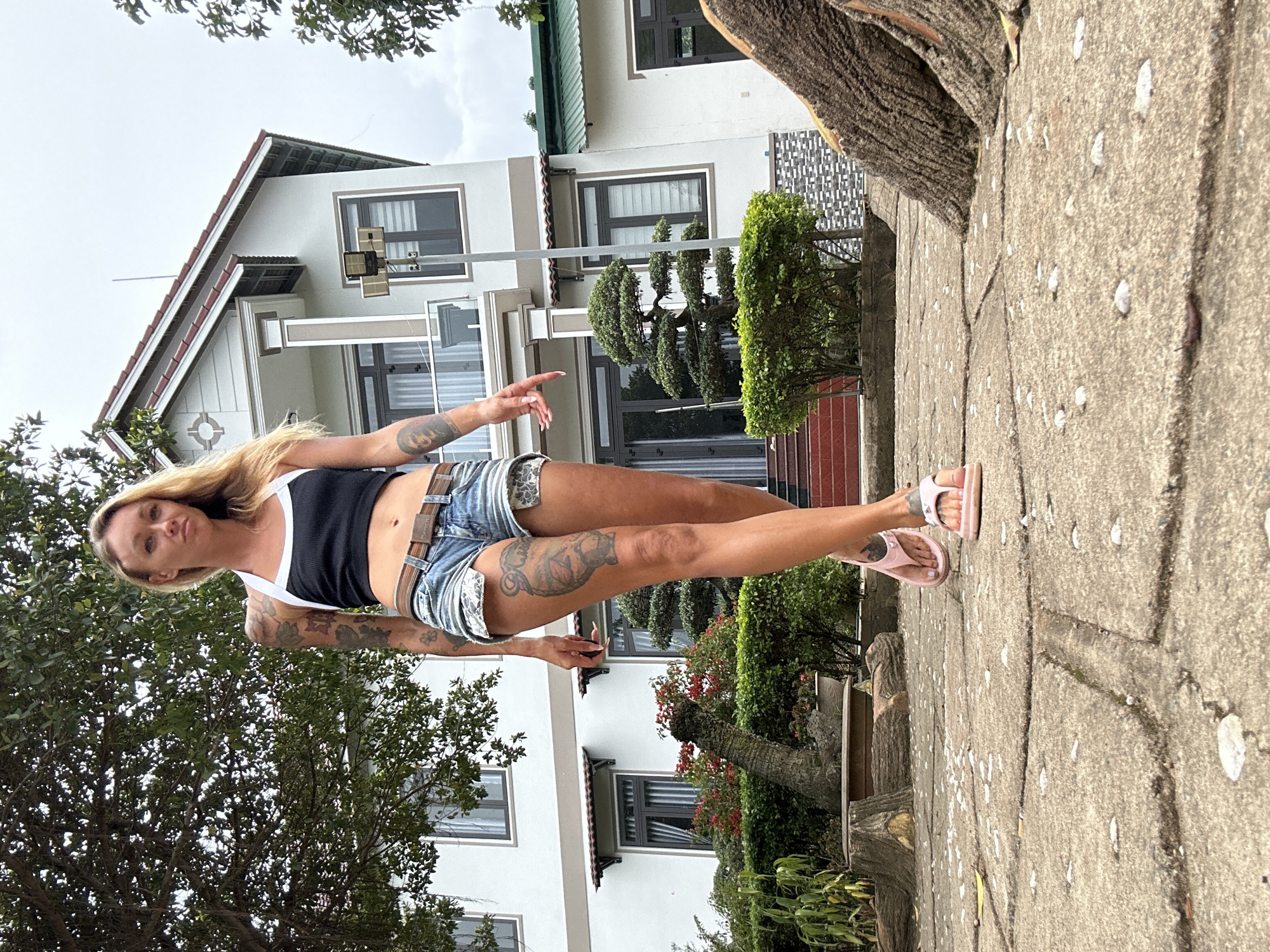 A woman with blonde hair and tattoos walks towards the camera on a concrete pathway in front of a modern white house. She wears a black crop top, denim shorts, and pink flip-flops. The house has multiple levels, dark-rimmed windows, and a green roof on one section. The pathway is made of large concrete slabs with debris scattered on it. In the background, there are trees, bushes, and potted plants, including a bonsai tree. The scene appears to be in a residential area, possibly in Thôn 2, Vietnam, given the architecture and vegetation. The sky is overcast.