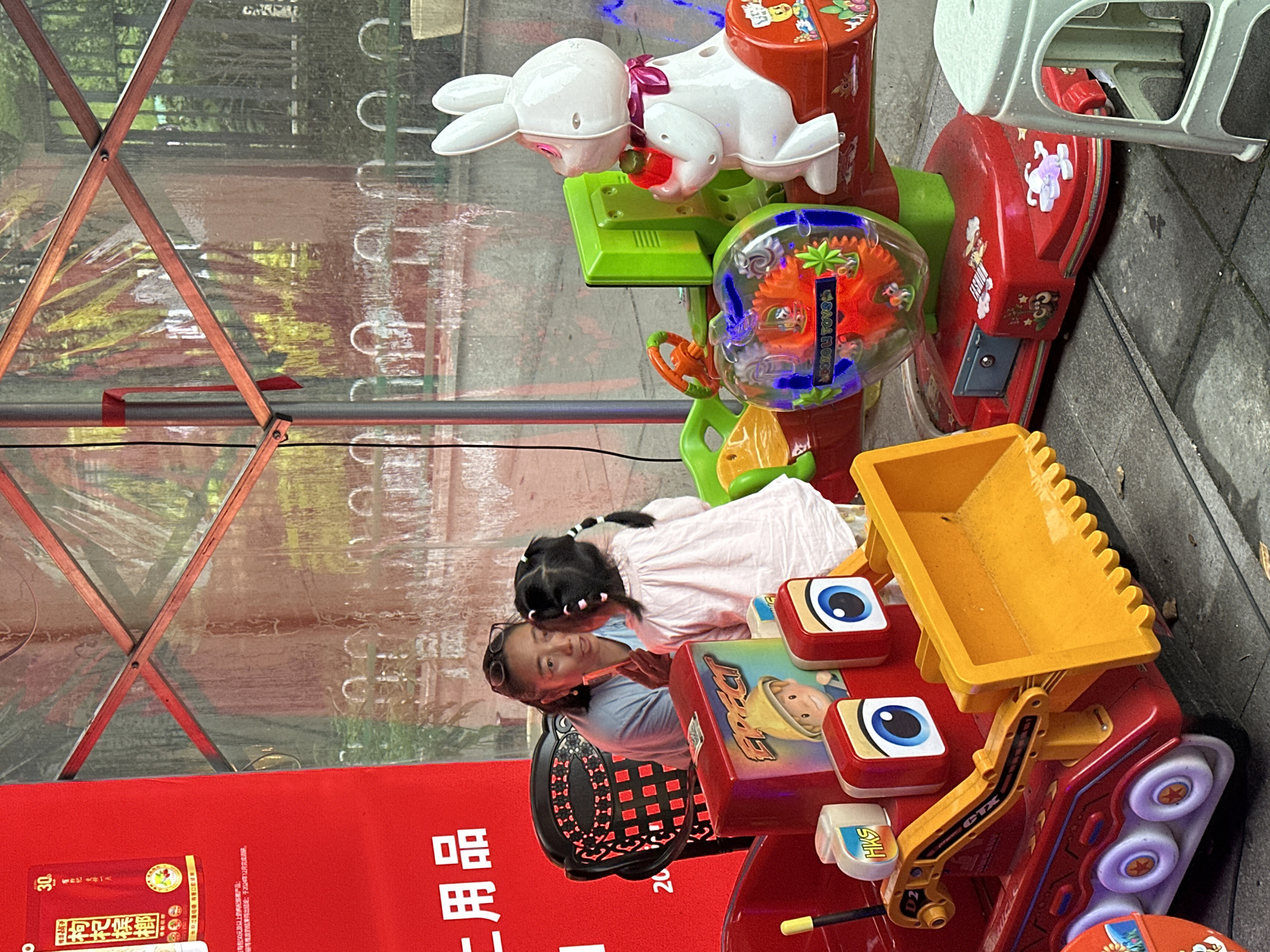 In the Chongqing High-tech Zone, China, a woman is seated in a red and black patterned chair, engaging with a young child. The child, facing away from the camera, wears a light pink top and has dark hair styled in pigtails with pink accents. The woman, with dark hair and sunglasses perched on her head, appears to be speaking to the child, holding a small red object. They are positioned behind a red toy construction vehicle with a yellow shovel and large blue eyes. To the right of the construction vehicle is a red and green kiddie ride with a white bunny figure. The background features a red banner with Chinese characters and a transparent tent structure with visible wooden supports and foliage reflecting on the transparent material. The ground is paved with grey tiles.