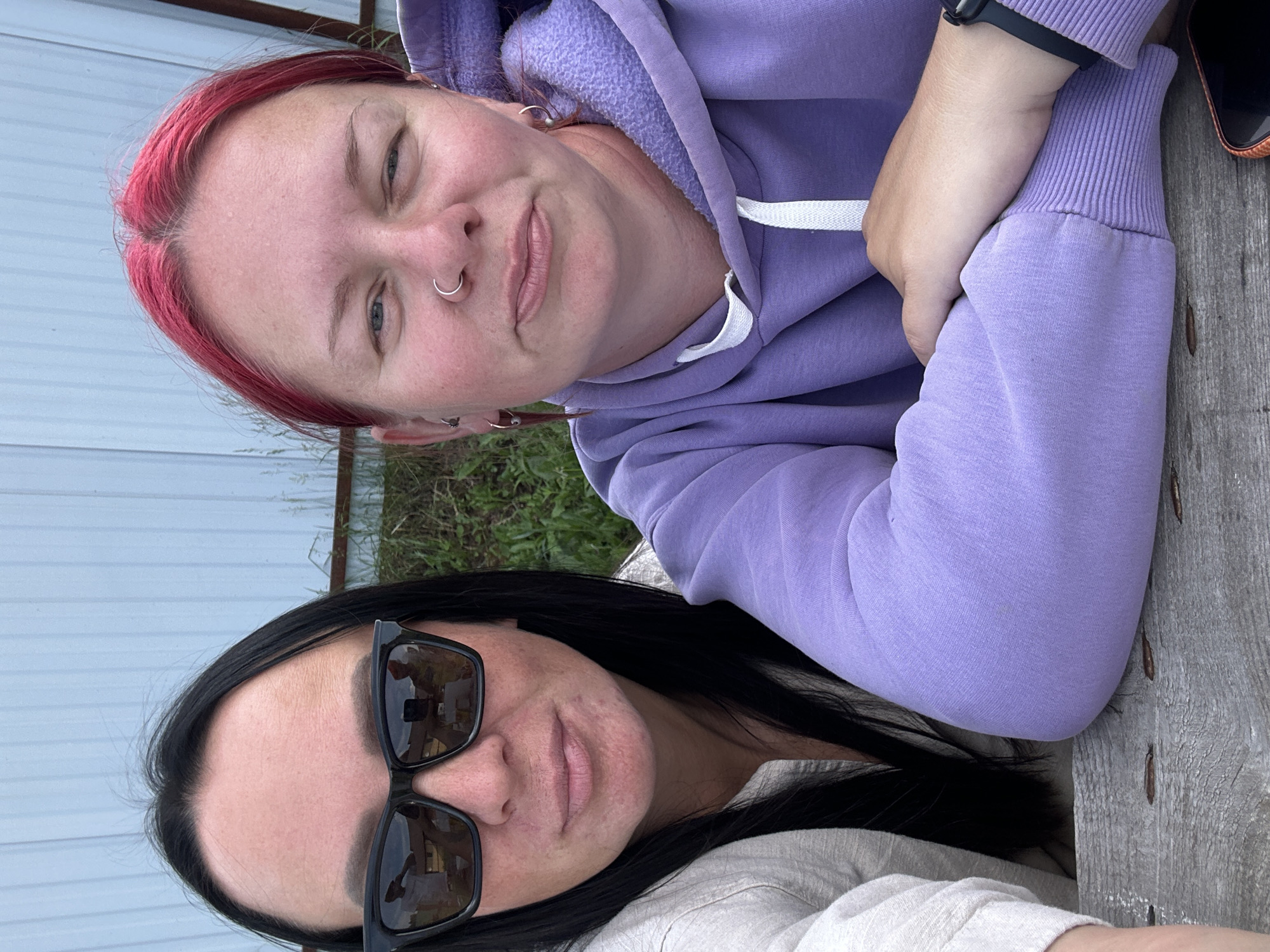 Two individuals are seated at a wooden table outdoors. The person on the left has long black hair and is wearing sunglasses and a light-colored long-sleeved shirt. The person on the right has bright pink hair, a nose ring, and is wearing a lavender hoodie. Their arms are resting on the table. In the background, there is a light blue structure with vertical lines and some greenery.
