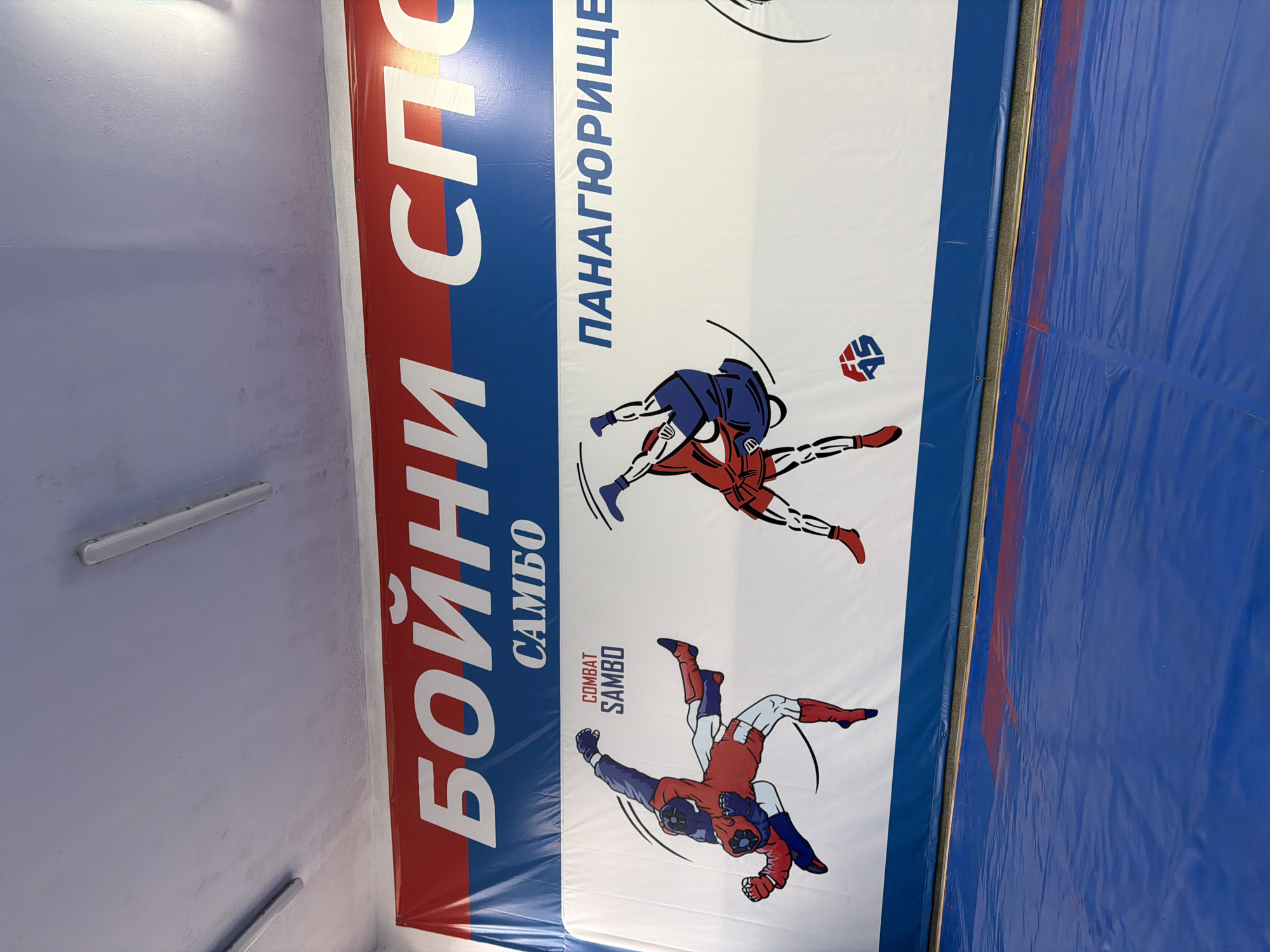 The image displays a brightly lit indoor sports facility, likely a martial arts or combat sports training area, seen from a slightly elevated perspective. The primary focus is a large white and blue padded banner or mat, positioned vertically on the left side of the frame. This banner features bold Cyrillic text and graphic illustrations of two stylized combatants.
The banner's design includes a red and blue strip on the left side, where a large white Cyrillic inscription "БОЙНИ СПО" (likely part of "Бойни Спортове" - Combat Sports) is visible. Below this, in smaller blue text, is "САМБО" (Sambo). Further down on the white section, the English text "COMBAT SAMBO" is printed in red. The banner prominently features two dynamic illustrations of Sambo practitioners in action, one wearing primarily red and blue attire, and the other predominantly blue with red accents, engaged in a grappling or throwing maneuver. Near the upper figure, the Cyrillic word "ПАНАГЮРИЩЕ" (Panagyurishte) is written in blue, indicating the location in Panagyurishte, Bulgaria. A small circular logo, possibly "SA," is also visible near the upper combatant.
To the right of the banner, a plain blue padded wall or mat extends across the rest of the frame, suggesting a safe training environment. The leftmost section of the image shows a light-colored, textured wall or ceiling with two white fluorescent light fixtures mounted horizontally. The overall impression is that of a dedicated space for Sambo training, emphasizing both sport and combat aspects.