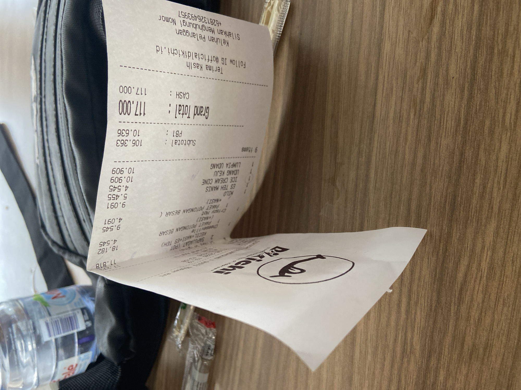 A folded receipt is placed on a wooden table. The receipt appears to be from a restaurant named "Dikichi". Some of the items listed on the receipt include "ES TEH MANIS", "ICE CREAM CONE", "UDANG KEJU", and "LUMPIA UDANG". The total amount is "117.000". There is also a bottle of water and a black bag in the background. The receipt has a logo with a drawing of a stylized face. The location is not specified in the image.