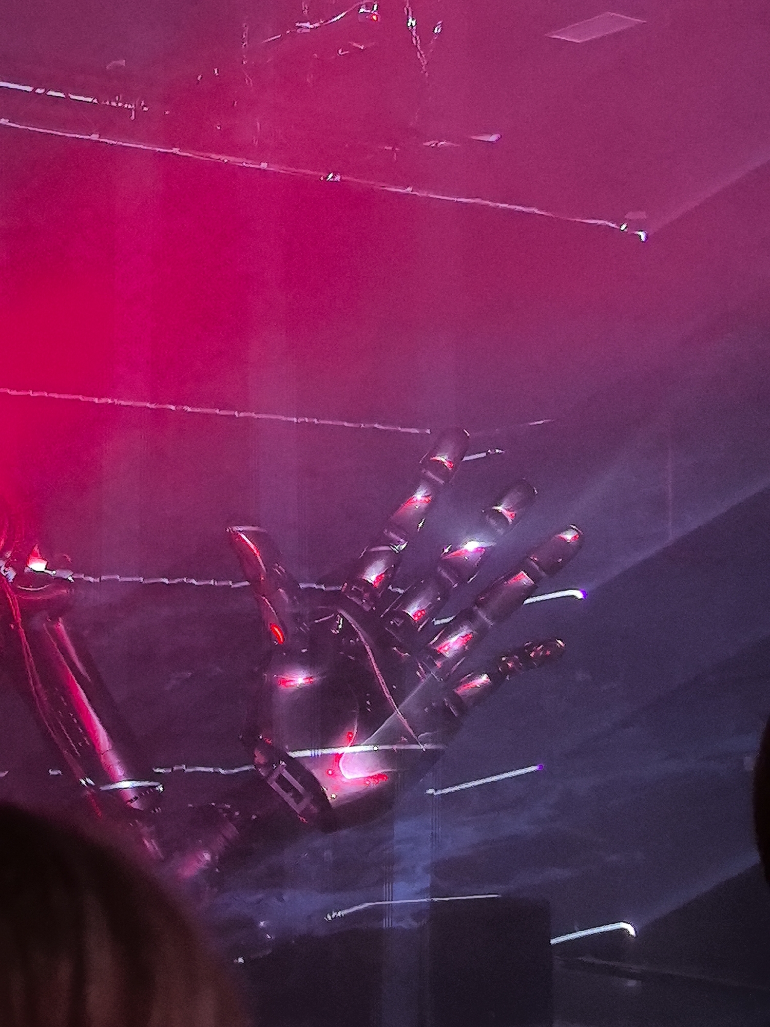 The image depicts a dimly lit, atmospheric scene dominated by vibrant pink and purple lighting. The central focus is a robotic hand, intricately detailed with metallic segments and illuminated by red lights within its joints. The hand appears to be posed in a gesture, possibly reaching out or making a statement.
Behind the robotic hand, a fine, almost web-like structure of cables or wires stretches across the frame, contributing to a slightly industrial or futuristic aesthetic. The background is shrouded in a hazy, smoky atmosphere, through which beams of light pierce the darkness, adding a dramatic effect. One prominent beam of light cuts diagonally across the lower right portion of the image.
On the left side, parts of a larger, also metallic structure are visible, potentially part of the same robotic system or stage machinery, bathed in a strong pink light. The overall impression is one of a stage performance, art installation, or a scene from a science fiction-themed event.
There are no visible people or recognizable landmarks that would definitively place this scene in the city of Perm, Russia, or any specific urban location. The environment is abstract and focused on the visual impact of light, shadow, and the mechanical form of the hand.