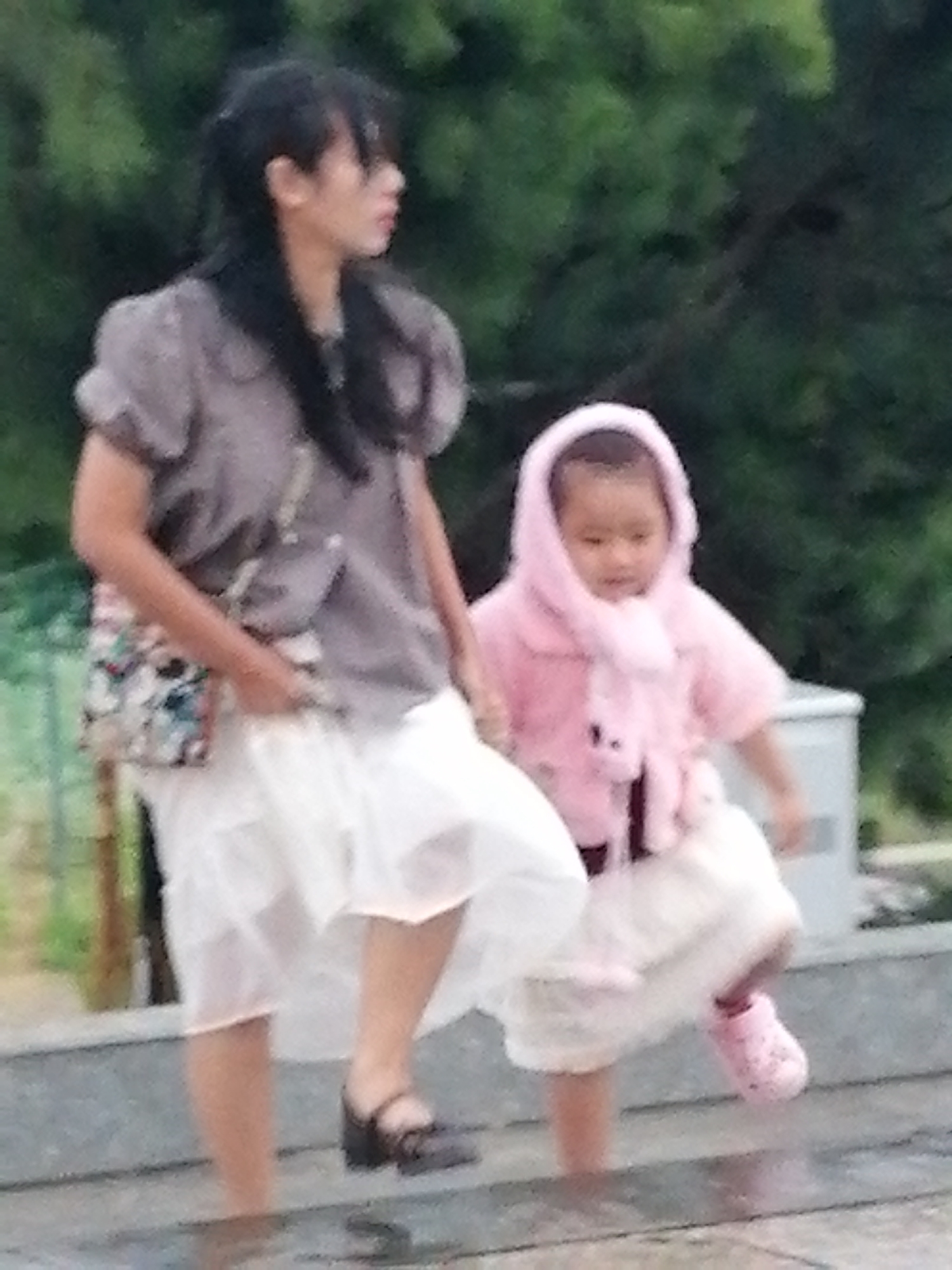 This is a photograph of a woman and a young child outdoors, possibly in Da Lat, Vietnam, given the green foliage which is characteristic of the region. The woman is standing on a raised platform or steps, wearing a grey short-sleeved top and a white, flowing skirt. She has dark hair and is holding a patterned purse. She appears to be looking to her right.
Beside her, the child is wearing a pink hooded jacket with a fuzzy trim and has pink shoes. The child is also on the steps, looking downwards. The background consists of blurred green foliage and a light-colored structure, possibly a bin or a wall. The overall image quality is low, with a grainy appearance. The lighting suggests it is daytime, likely overcast or in a shaded area.