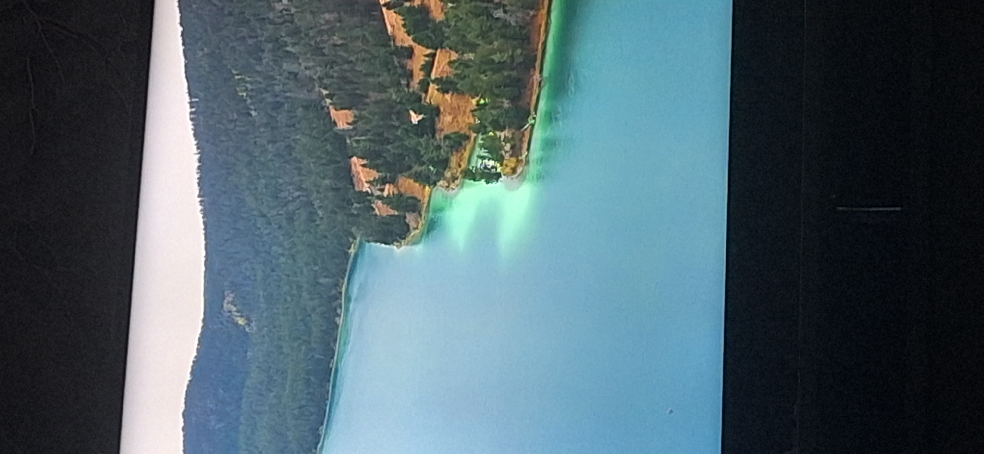 The image shows an aerial view of a serene lake surrounded by lush green forests and rolling hills. The water is a brilliant turquoise, with lighter patches indicating shallow areas or underwater sandbars. The shoreline is dotted with dense evergreen trees, and on the right side of the frame, a clearing on a hill reveals a small cluster of buildings with orange roofs, suggesting a settlement or resort. The overall impression is one of natural beauty and tranquility, typical of a mountain or lake landscape. However, this image does not depict the city of Bila Tserkva, Ukraine, as the scenery is mountainous and forested, unlike the flatter terrain of that city.