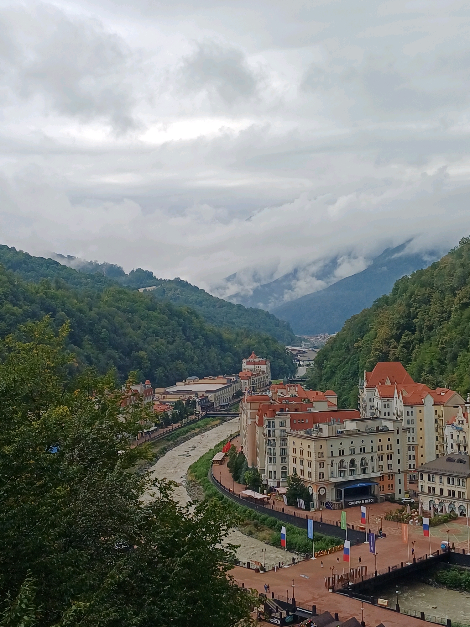 The image captures a panoramic view of Estosadok, Russia, a resort town nestled within a valley. The scene is dominated by lush green mountains that rise on either side of a winding river. The river, a muddy brown, flows through the center of the frame, bordered by a pedestrian walkway and several buildings.
The architecture of these buildings is reminiscent of European alpine villages, with pastel facades, red-tiled roofs, and multiple stories. Some of these buildings appear to be hotels or resorts, with balconies and large windows. In the foreground, a cluster of these buildings lines the riverbank, with a public square featuring flags and a stage. The stage has text in Russian, which translates to "Watch in Summer."
The sky is overcast with thick, grey clouds, and pockets of fog or mist are visible clinging to the mountain peaks, adding a dramatic atmosphere to the scene. There are a few scattered people visible on the walkway and in the square, but no significant activity is apparent. The overall impression is one of a scenic, somewhat melancholic, mountain resort town under a cloudy sky.