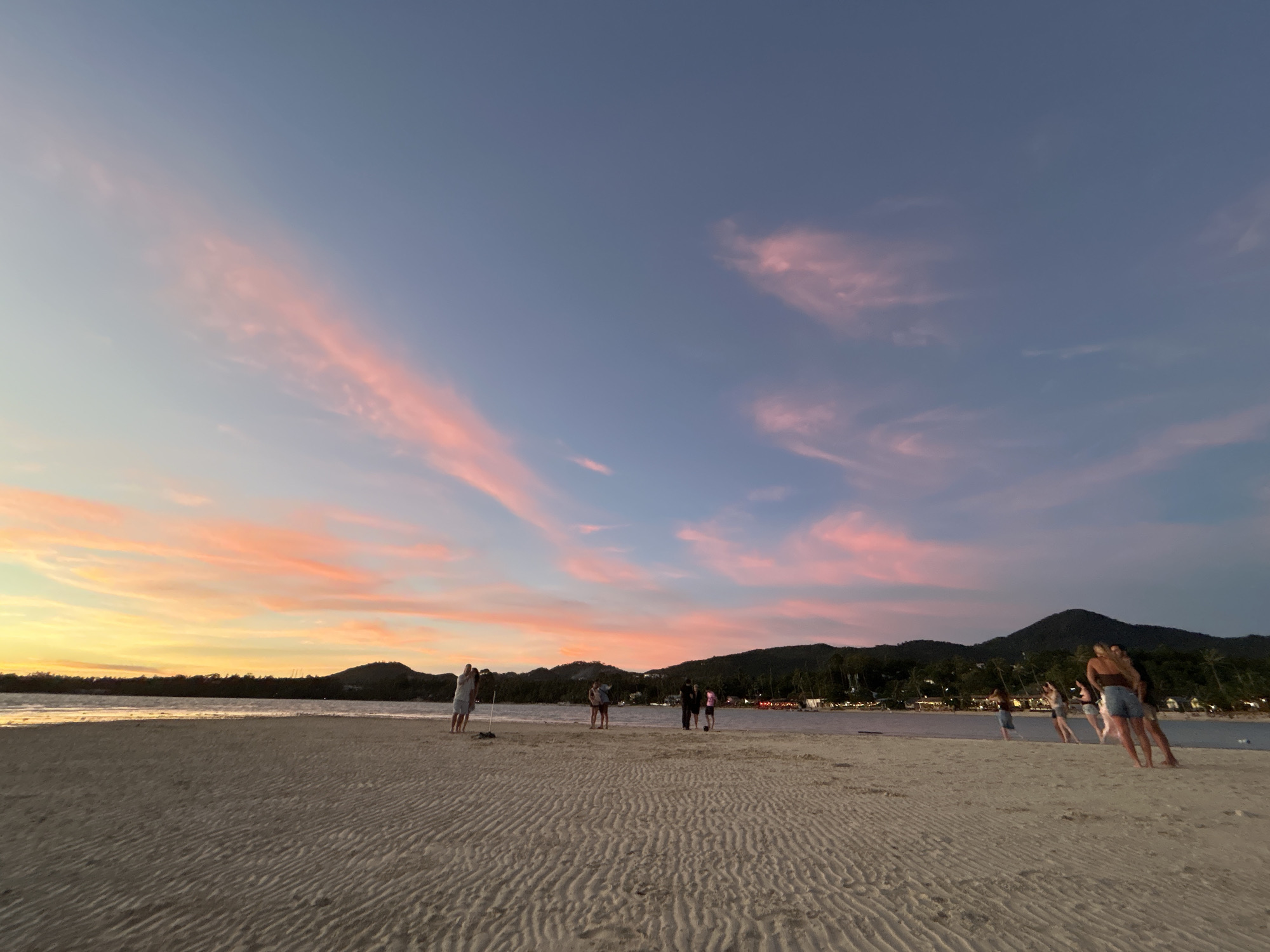 This image captures a serene sunset scene on a sandy beach, likely located in ตำบลเกาะพะงัน, Thailand. The sky is a canvas of gradient hues, transitioning from a warm yellow and orange near the horizon to soft pinks and purples in the clouds, eventually deepening into a dusky blue overhead. The textured sand in the foreground, marked with wave-like ripples, suggests the receding tide. A line of lush, green hills forms the backdrop, dotted with trees and illuminated by scattered lights that hint at a settlement or resort. Several individuals are visible on the beach. On the left, a couple stands close together. Further to the right, a group of four people are gathered, appearing to interact or pose for a photo. In the far right of the frame, two women are walking away from the camera. The overall atmosphere is tranquil and picturesque, indicative of a popular tourist destination during a beautiful twilight.