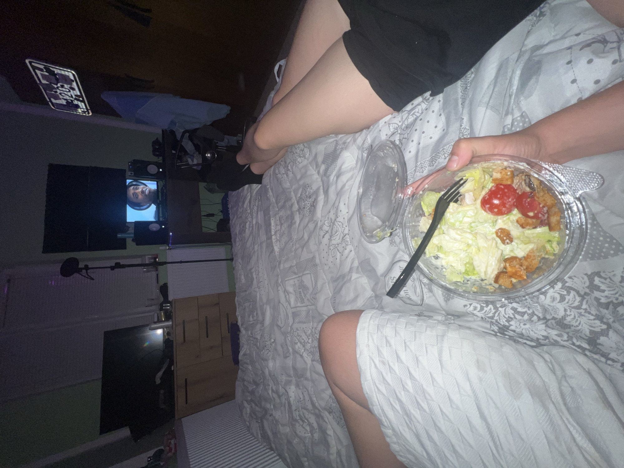 A person is in bed eating a salad. The person is wearing dark shorts and a light-colored garment. The salad is in a clear plastic container with a black fork. The bed has a patterned white and gray comforter. In the background, there is a TV showing a person wearing headphones, a microphone, a digital clock, and a window. There is no visible evidence suggesting that this image was taken in Trifonovo, Bulgaria.