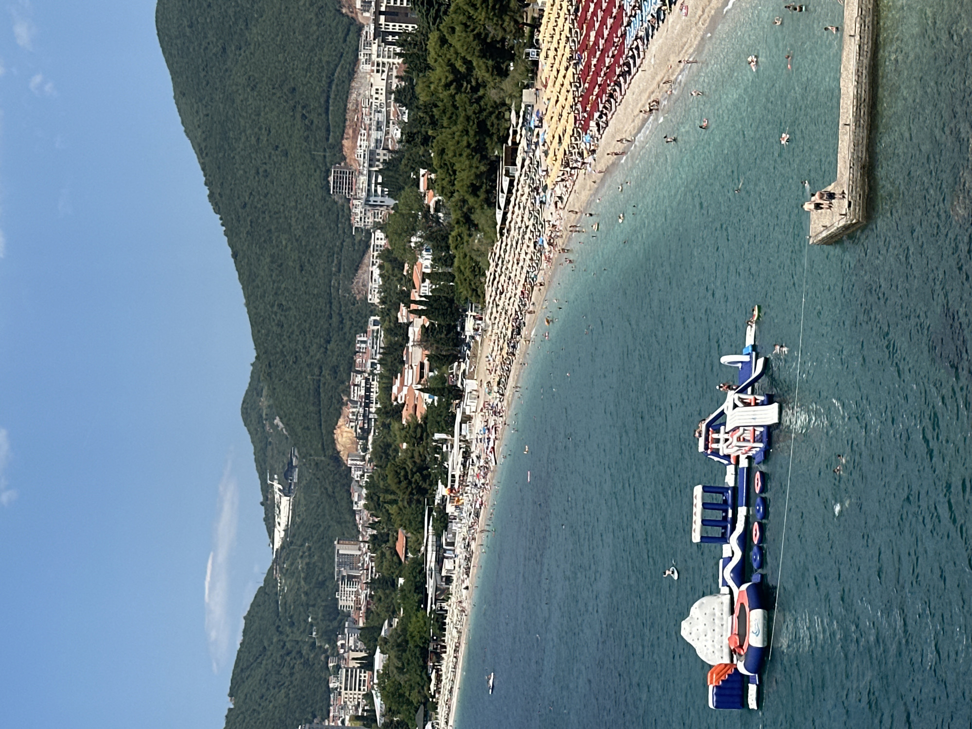 The image captures a picturesque view of a coastal city, identified as Budva, Montenegro, on a clear, sunny day. The foreground is dominated by the turquoise waters of the Adriatic Sea, where an inflatable water park with slides, climbing walls, and trampolines is set up. Several individuals are visible enjoying the water park and swimming in the sea.
To the right of the water, a concrete pier juts out into the sea, with two men standing on it. The coastline features a long, sandy beach crowded with beachgoers relaxing under numerous yellow and red umbrellas. Behind the beach, a dense array of trees lines the shore, interspersed with buildings of varying architectural styles, indicative of a developed urban area.
In the background, lush green mountains rise, their slopes dotted with more buildings and structures, some appearing to be under construction. The sky is a bright blue with a few wispy clouds, suggesting pleasant weather conditions. The overall scene depicts a vibrant summer day at a popular seaside resort.