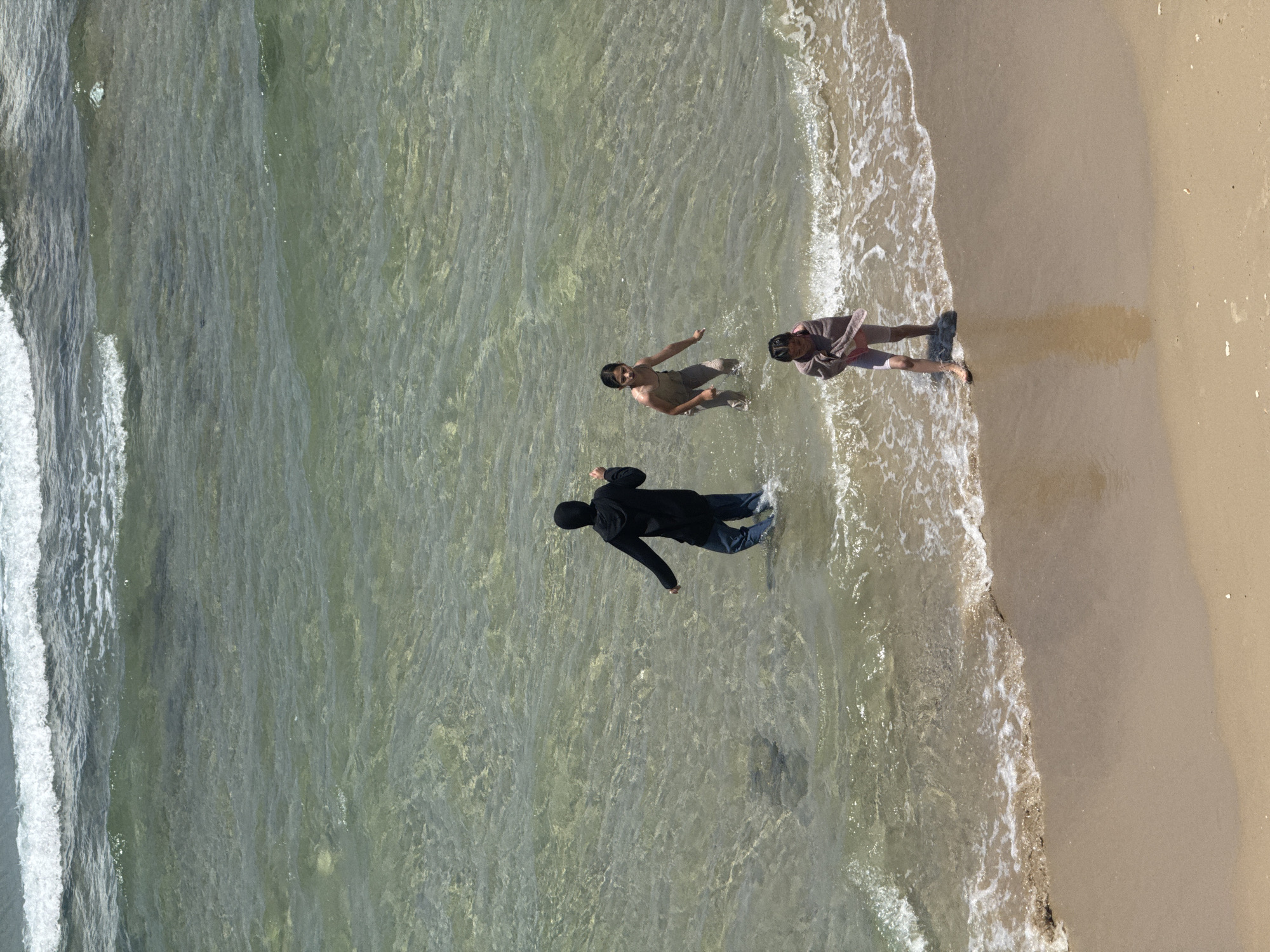 The image shows three individuals at the beach in Tel-Aviv, Israel. One person wearing a black long-sleeve shirt and dark pants is standing in the shallow water. Another individual in light-colored clothing is also standing in the water with their arms outstretched. The third person is standing on the beach near the water's edge, wearing a long-sleeve shirt and shorts. The scene is an overhead shot, showing the water, waves, sand, and people.