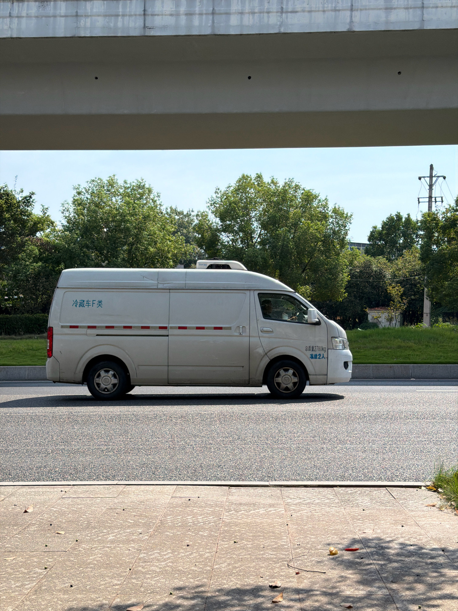 A white refrigerated van is driving on a road in the city of Jiangjin, China. The van has blue text on its side. The van is driving under a large overpass. Trees and a utility pole are visible in the background. The sky is blue with some clouds. The road is asphalt and there is a sidewalk to the right of the road. The sidewalk is paved with concrete and has some fallen leaves on it. The lighting suggests it is daytime.