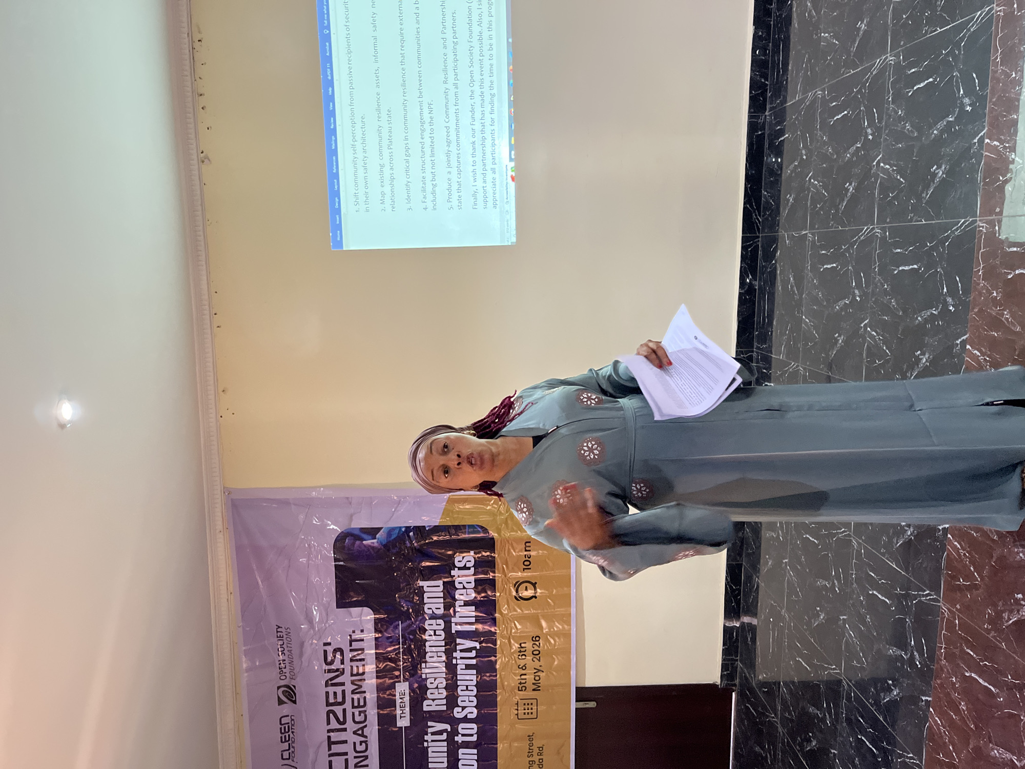 A presentation or meeting is taking place in an indoor setting. The speaker, a woman with auburn braids and wearing a teal-colored dress with embroidered detailing, is holding papers and gesturing with her hand. Behind her, there is a projector screen displaying text, likely part of her presentation, and a banner bearing the words "CITIZENS’ ENGAGEMENT" along with the theme: "Community Resilience and on to Security Threats". The banner also indicates the dates "5th & 6th May, 2026" and the time "10 am", as well as the location "ng Street, da Rd". The Open Society Foundations logo is present on the banner, suggesting they may be sponsors or partners of the event. The floor is tiled in a black and dark brown marble pattern. The city is Unknown, Unknown.