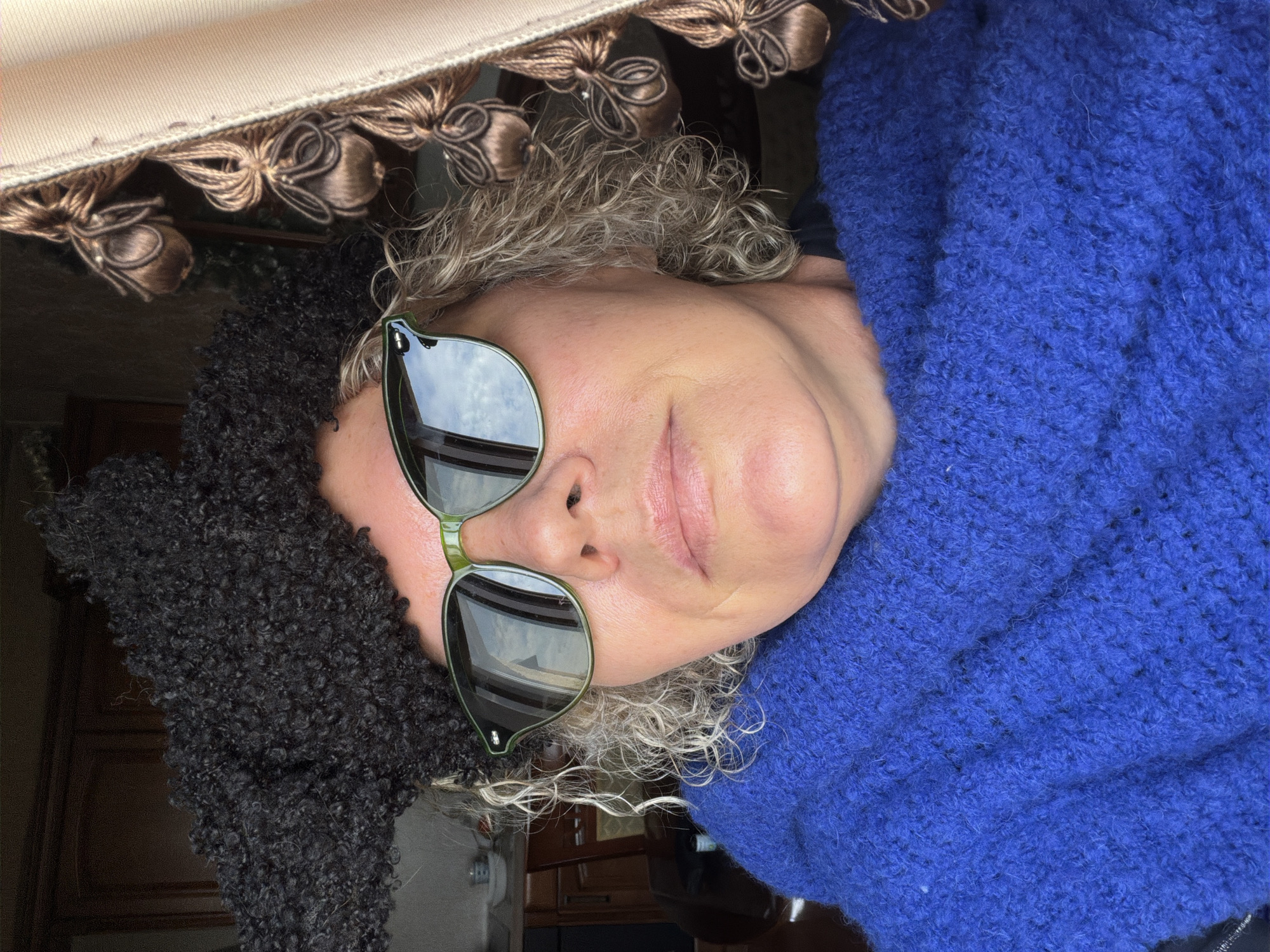 The image shows a close-up, selfie-style portrait of a person wearing a black, curly hat, green-rimmed sunglasses, and a bright blue, textured scarf or shawl. The person's wavy, light-colored hair is visible around their face and shoulders. The background is out of focus but suggests an indoor setting with cabinetry and a window with tasseled curtains. The image does not contain any specific visual cues that would allow for an identification of the location as Surgut, Russia.
