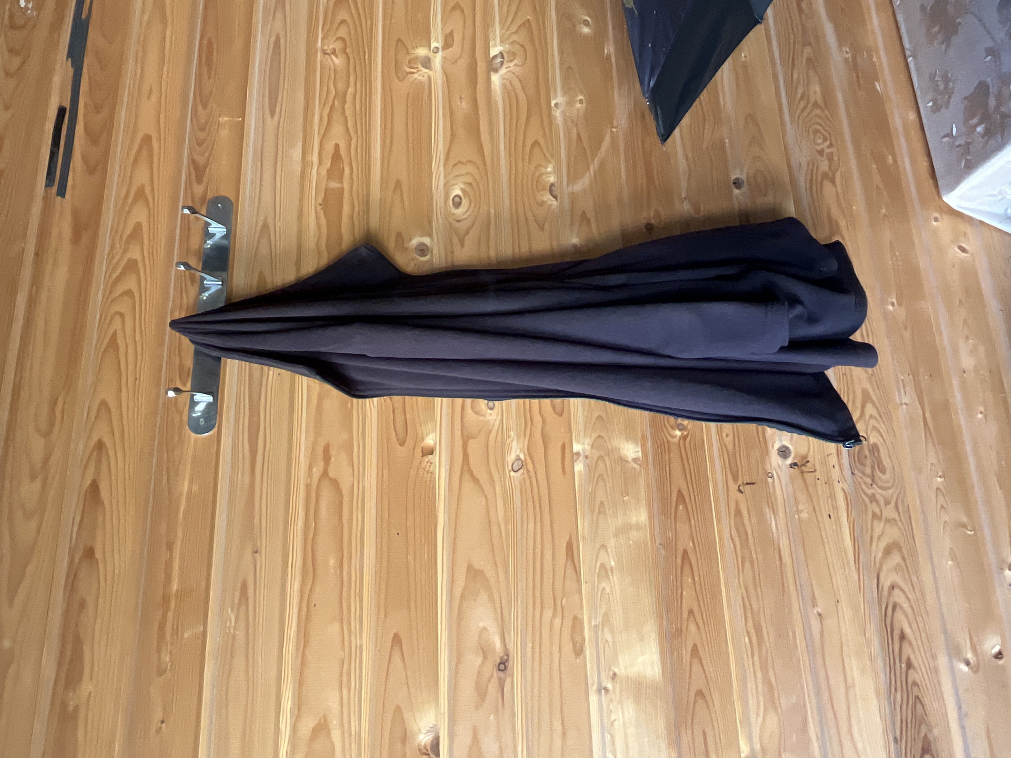 A dark garment, possibly a coat or robe, hangs from a metal rack with three hooks mounted on a wooden paneled wall. The garment appears to be made of a thick material and is gathered at the top. The wall consists of horizontal wooden planks with visible grain and knot patterns. To the right of the garment, a portion of a dark, flat surface, possibly a table or lid, is visible at the bottom right corner of the frame. There are no people or animals in the image. The scene is indoors, with the lighting suggesting it is daytime. The location is not specified within the image itself.