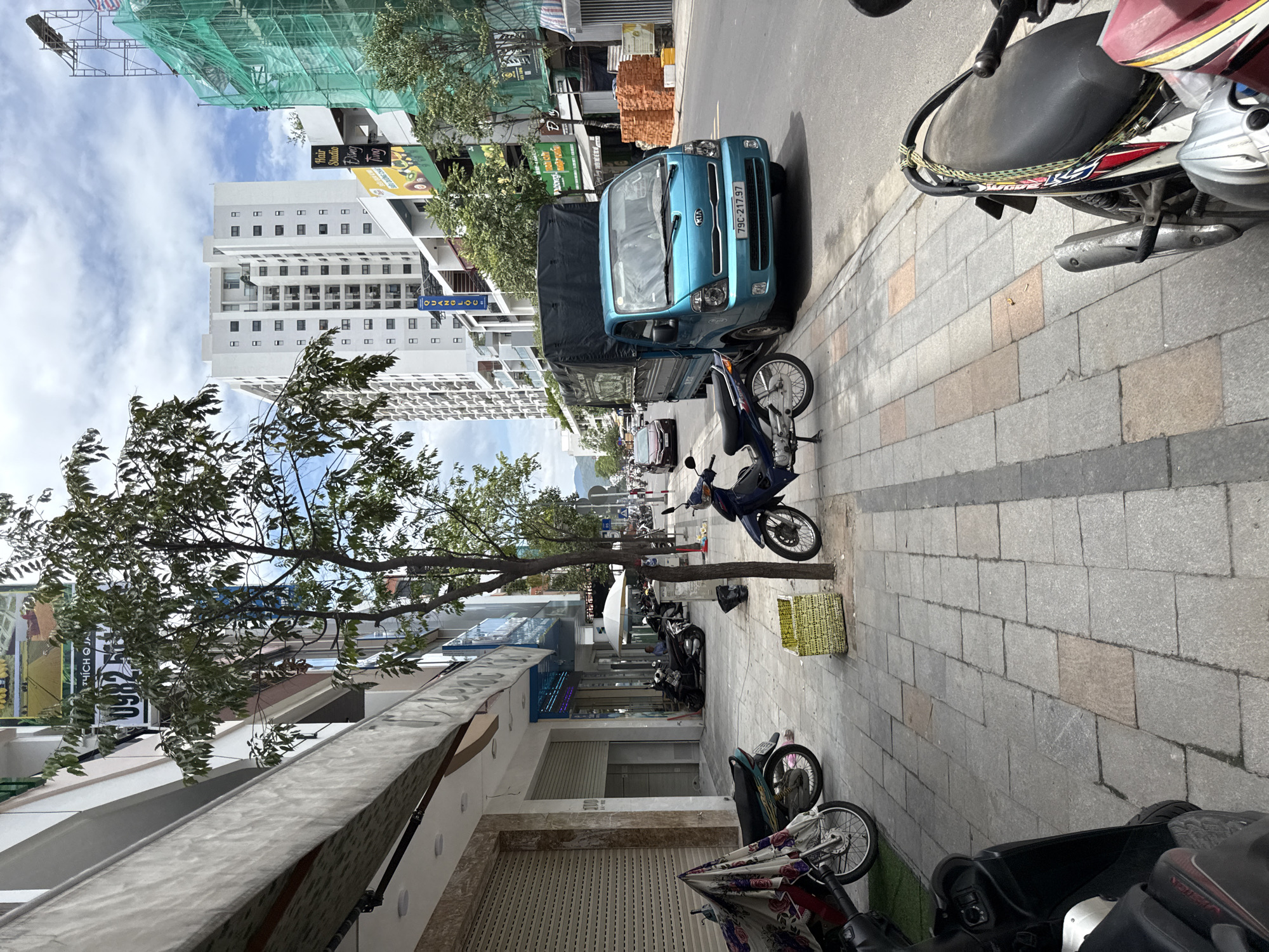 This image depicts a street scene in Nha Trang, Vietnam, likely on a commercial street with buildings lining one side and a road on the other. The foreground is dominated by a sidewalk paved with grey and tan stone tiles. Several motorbikes are parked along the sidewalk, some with patterned fabric draped over them. A bright turquoise Kia truck is parked on the road, facing right. Further down the street, a tall white apartment building stands out against a partly cloudy sky, with scaffolding and green netting visible on a building to its right, indicating ongoing construction. The street appears to be moderately busy, with other vehicles visible in the distance. Trees are planted along the sidewalk, providing some shade. The buildings on the left have storefronts with rolling metal shutters, suggesting they may be closed or about to open.