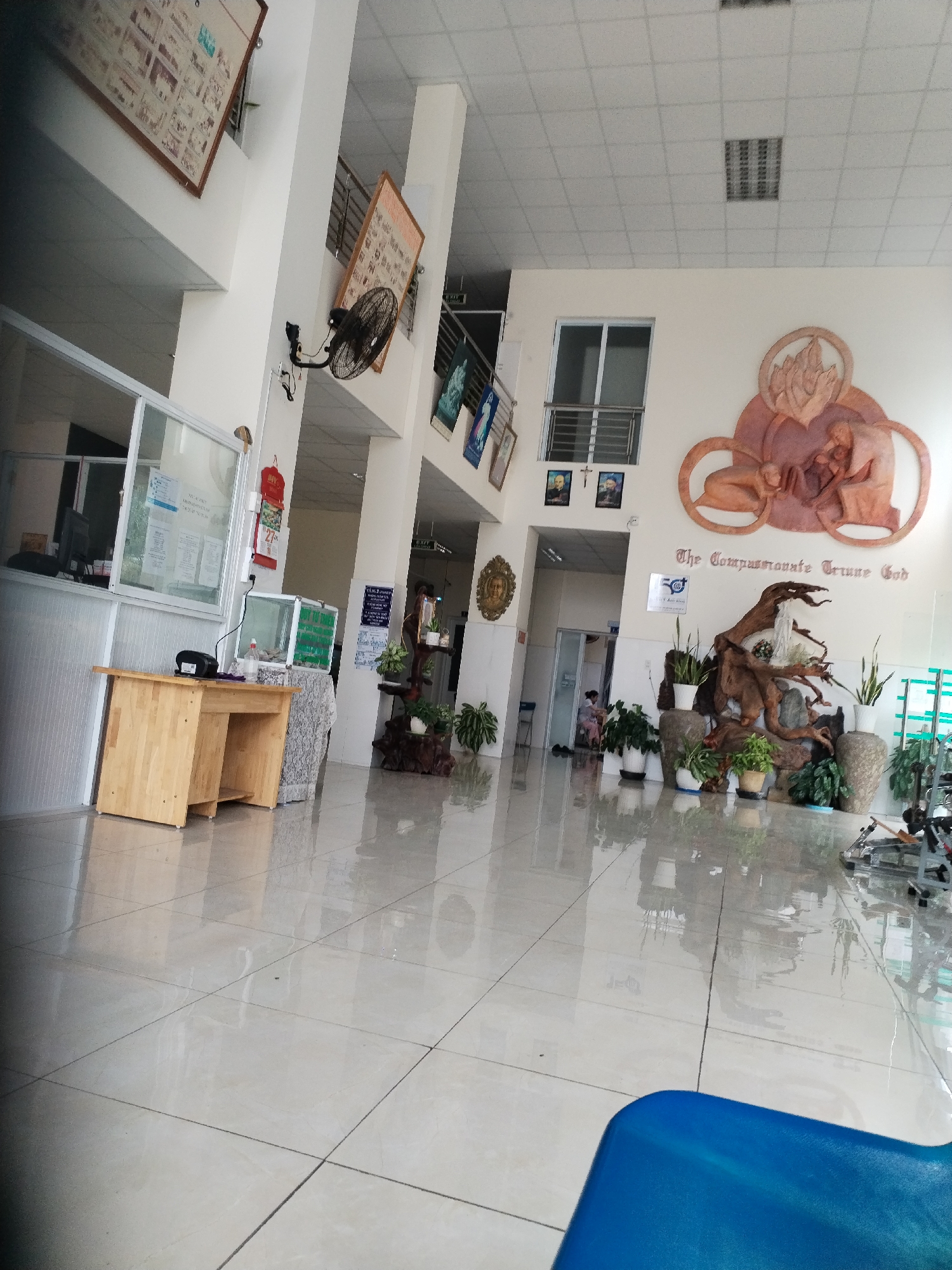 The image shows the interior of a building in Nam Nha Trang Ward, Vietnam, likely a hospital or clinic waiting area. The floor is tiled and highly polished, reflecting the interior light. There is a reception desk to the left with a wooden counter. Further into the space, there's a seating area and several potted plants arranged around a decorative driftwood sculpture. A large artwork depicting figures, possibly religious in nature, is visible on the far wall along with smaller framed pictures. A person in a medical gown is walking through a distant doorway. A blue plastic chair is in the foreground. Artwork and informational posters are displayed on the walls. A ceiling fan is mounted on one of the columns.