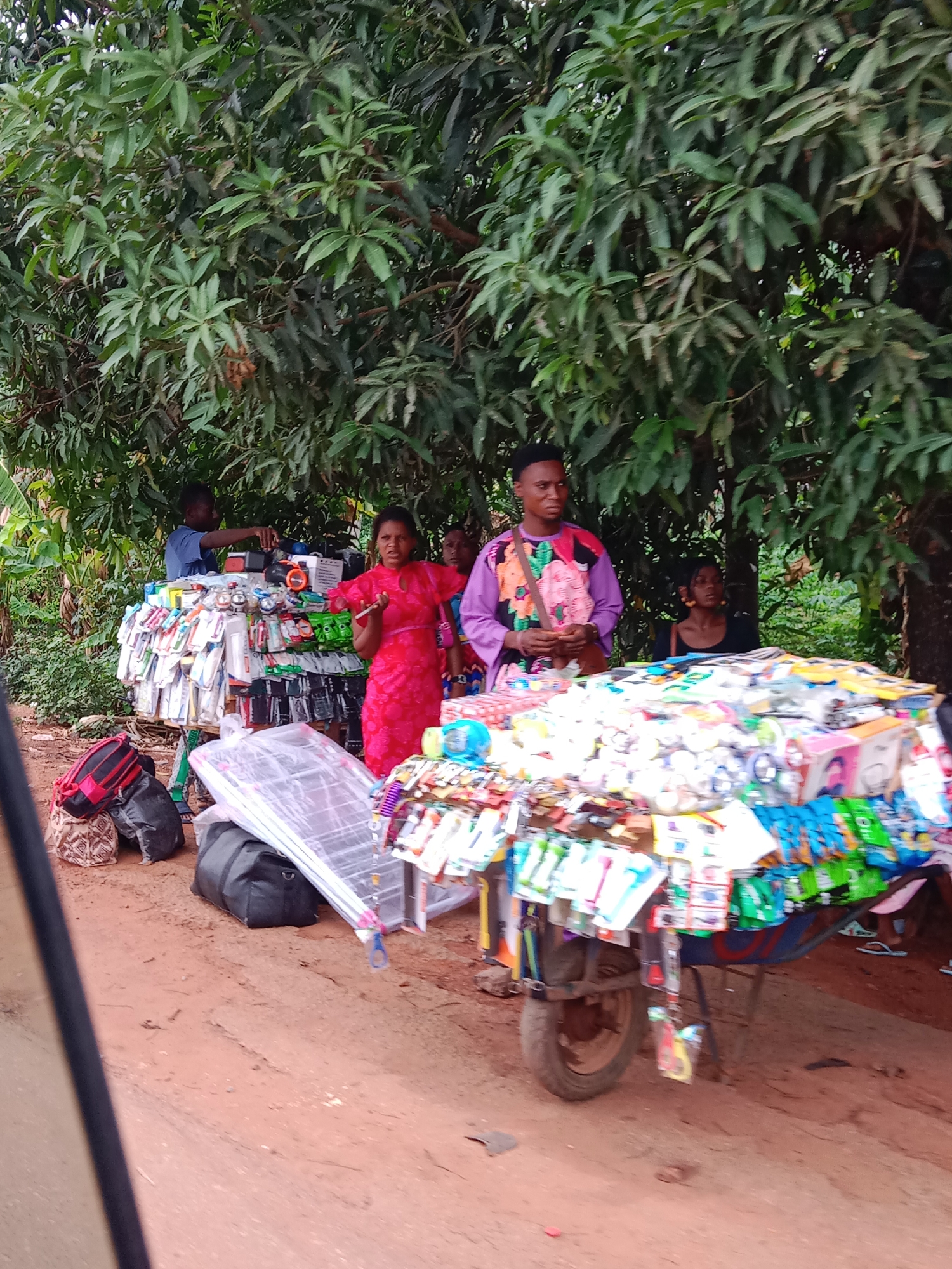 The image shows a roadside vending scene in an unknown city. Several individuals are gathered around two makeshift stands or carts, heavily laden with small merchandise items such as phone accessories, toiletries, and small toys. The environment is shaded by a large leafy tree. One person wears a pink dress and another a brightly patterned purple shirt. Luggage or large bags are situated near one of the carts. The ground appears to be a dirt road.