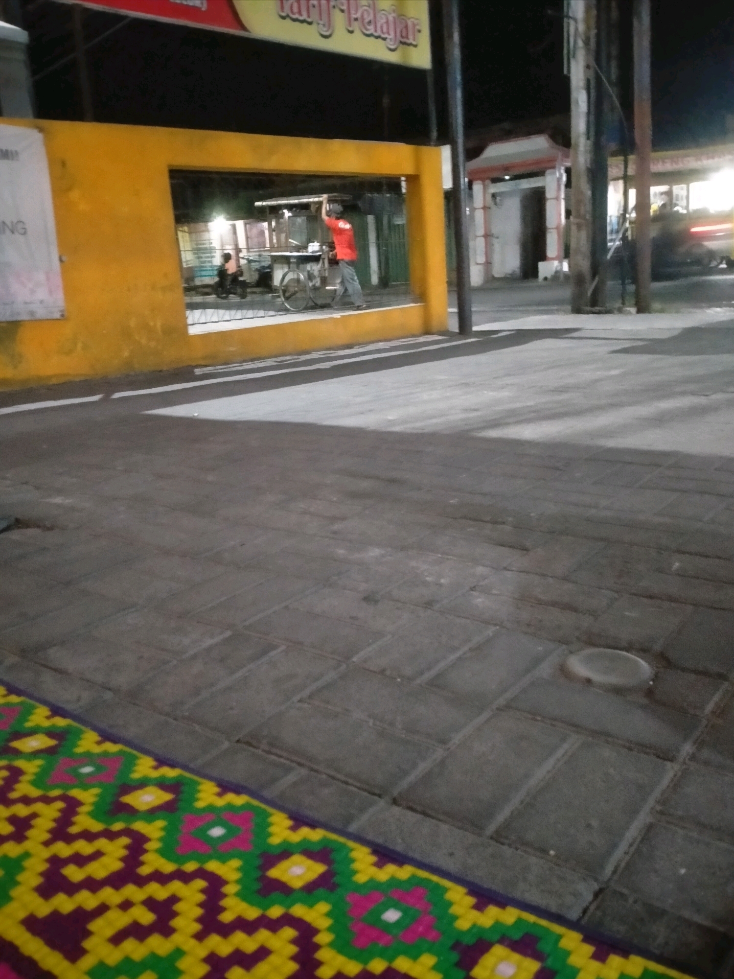 The image depicts a scene at night in the city of Kasihan, Indonesia, likely near a school or educational institution given the partially visible text on a sign that reads "SMU" and "Pelajar" (student). In the foreground, a patterned mat in yellow, green, pink, and purple is laid on a paved walkway. The walkway is made of grey rectangular bricks. Beyond the walkway, a yellow structure with an opening is visible. Through the opening, a man in a red shirt is standing next to a cart with a bicycle wheel. He appears to be engaged in some activity, possibly related to selling food or goods. Another person on a scooter is also visible inside the opening. The background shows a street with poles and some blurred lights, indicating it is nighttime. The overall impression is of an outdoor scene at dusk or night in an urban environment.