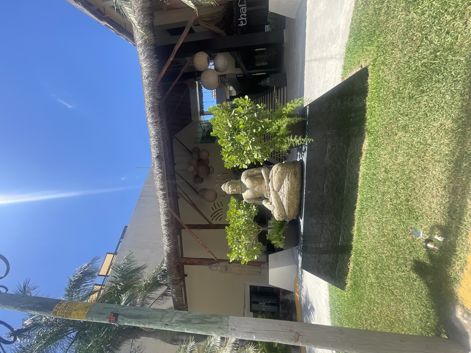 The image captures the entrance of the Thai restaurant "Thai Way" in Playa del Carmen, Mexico. The restaurant's facade features a traditional thatched roof with hanging wicker lamps. A stone Buddha statue sits amongst greenery at the entrance, flanked by a black stone platform and adjacent lawn. A utility pole is visible in the foreground. The sky is clear and blue.