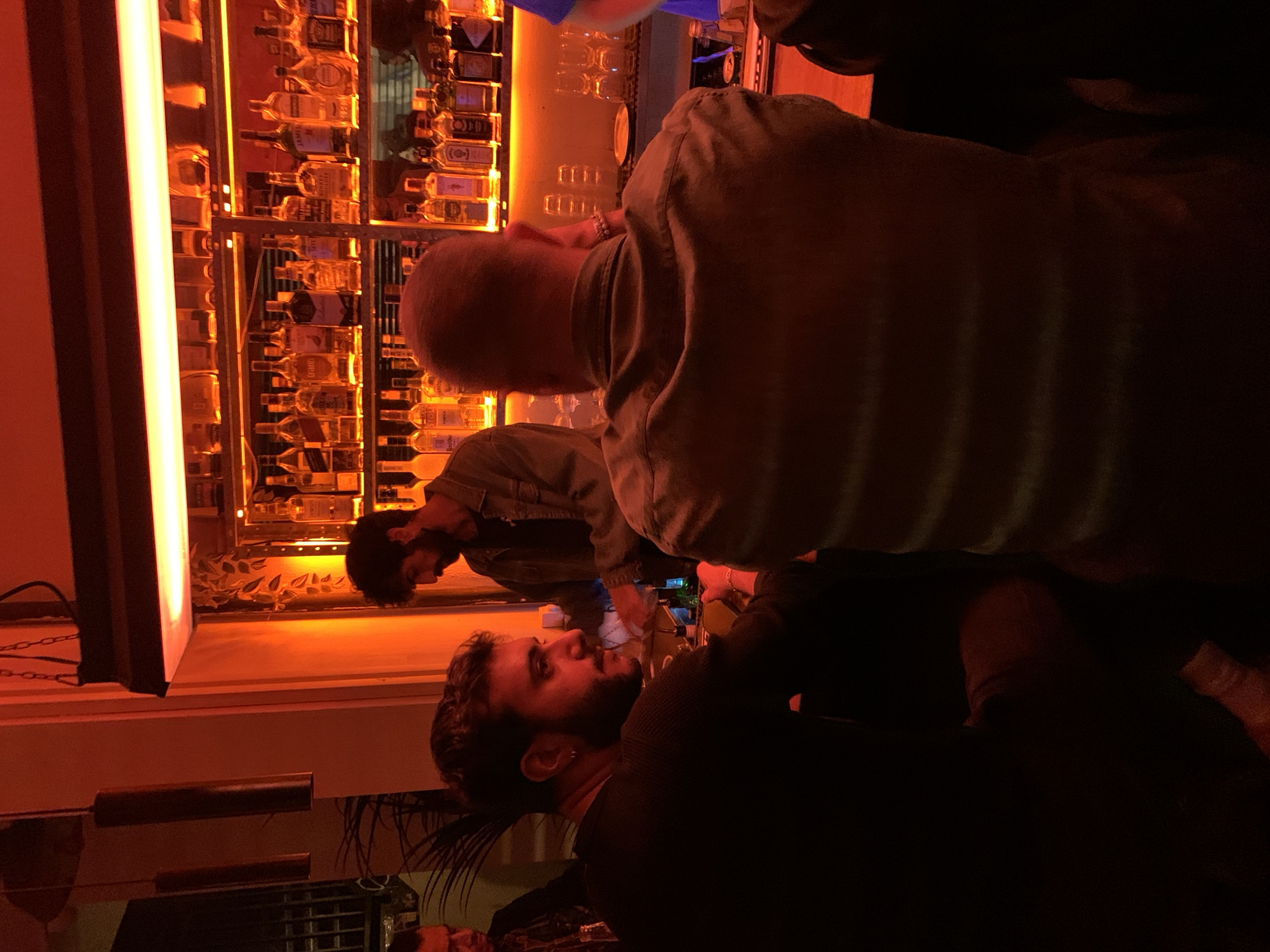 The image shows three men at a bar in Tel Aviv, Israel. The bar has a back-lit display of liquor bottles. One man is bald and wearing a khaki shirt. Another man has a beard and curly hair. The third man is partially visible. The scene is illuminated by orange and red lighting.