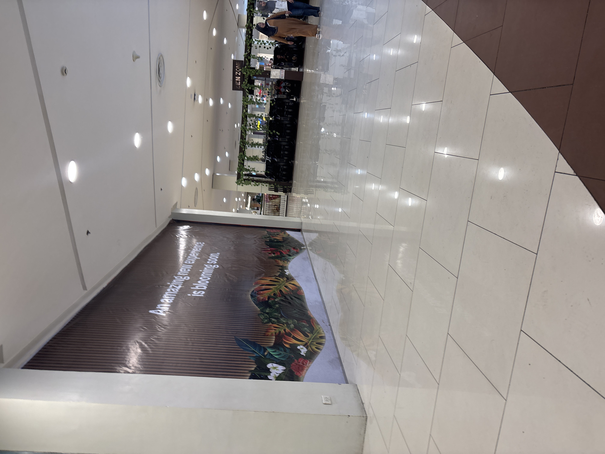 The image captures a brightly lit, tiled interior of a commercial space, likely a shopping mall, in Davao City, Philippines. A large banner stretches across the wall, adorned with a nature-themed graphic and the text "An amazing new experience is blooming soon." The floor is a polished, light-colored tile, reflecting the overhead lighting. In the background, several dark, massage chairs are visible, with a sign above reading "ZEN ZONE." Two women, one wearing a tan abaya and headscarf, and the other a white jacket and headscarf, are walking in the middle ground. The overall impression is one of a clean, modern retail environment.
