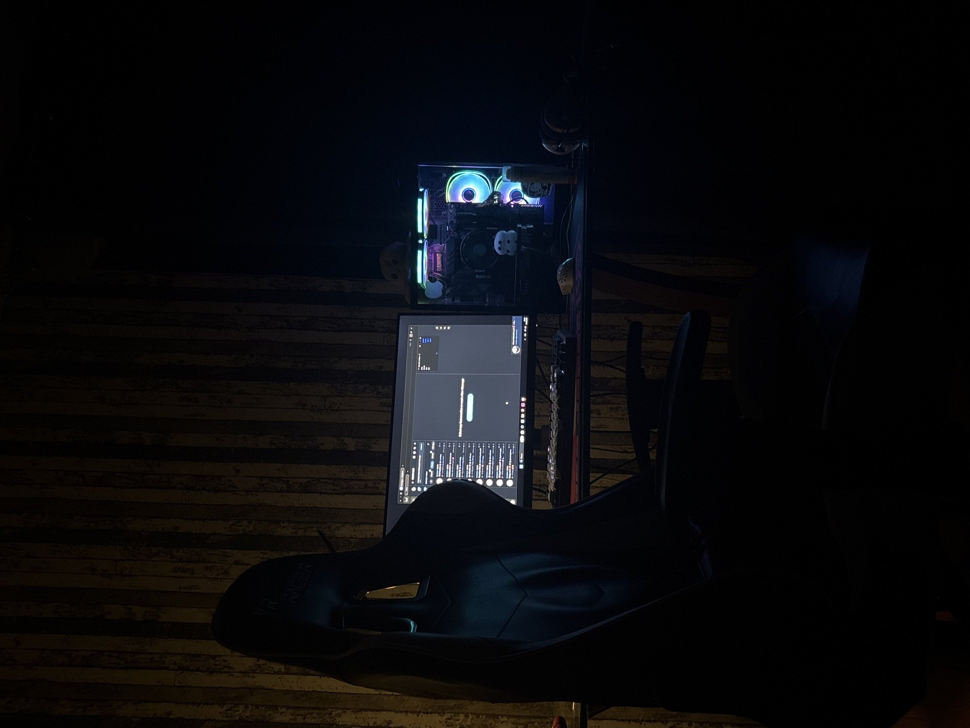 An indoor, low-light scene shows a gaming setup with a computer tower, monitor, and gaming chair. The computer tower has RGB lighting and is positioned on a desk. A keyboard and mouse are visible on the desk in front of the monitor. The gaming chair is black and appears to be made of leather or a similar material, with a high back. The background features a wall with vertical wooden paneling. The lighting in the room is dim, with the primary light sources being the RGB lighting from the computer and the monitor display. The overall impression is that of a dedicated gaming space. The location is described as Unknown, Unknown.